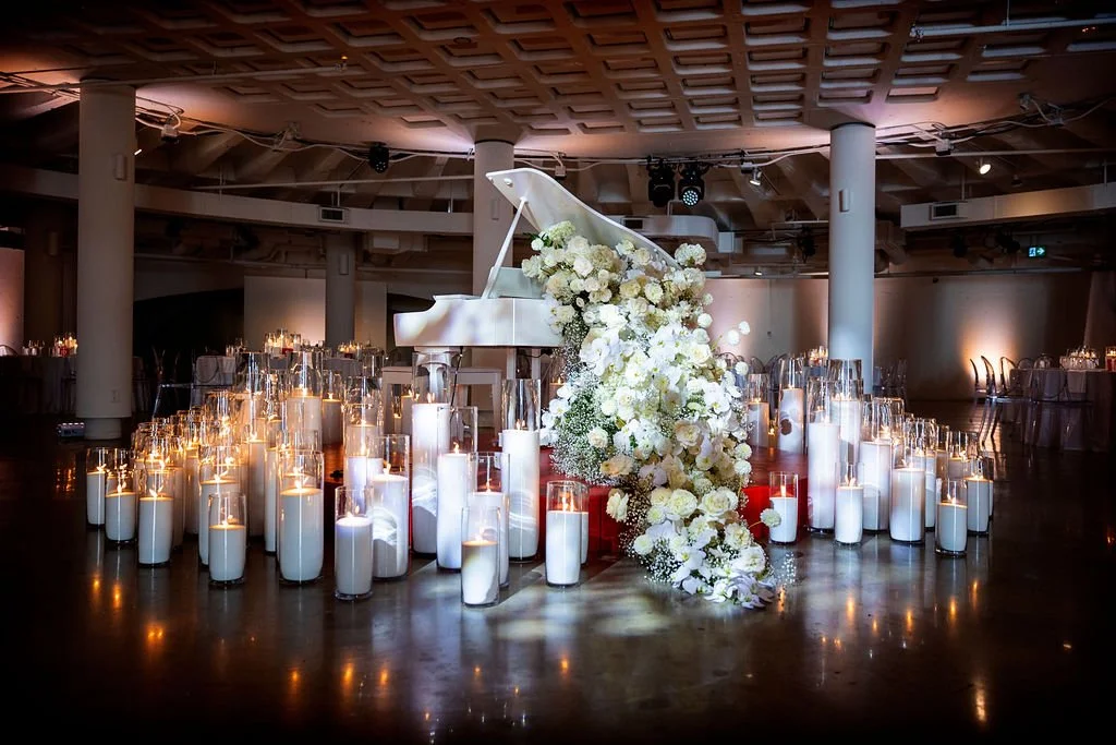A white grand piano decorated with white flowers and greenery, surrounded by tall white candles inside glass holders, in a dimly lit event hall with a high ceiling and round tables in the background.