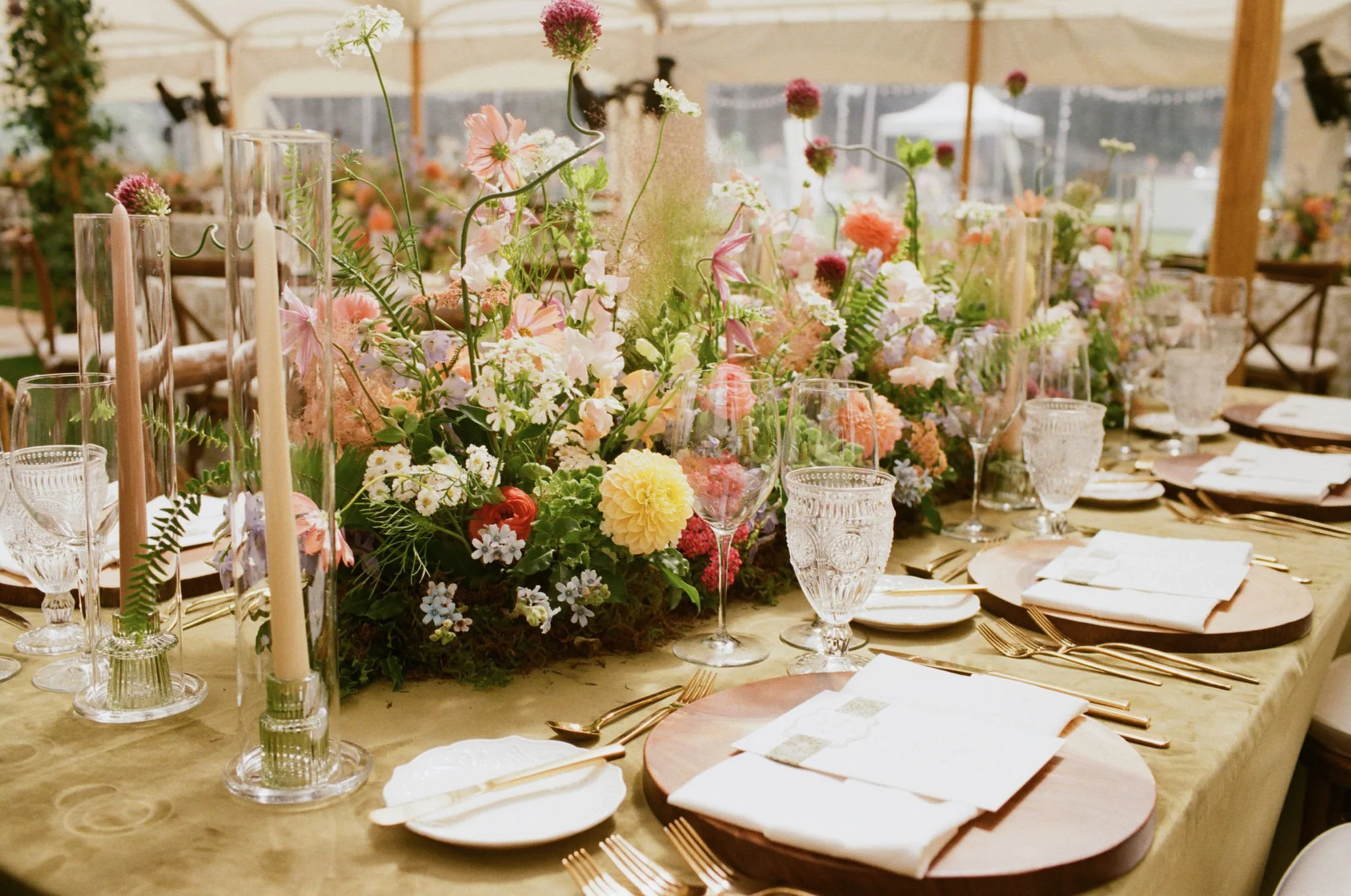 Elegant wedding table setting with a large floral centerpiece, candles, crystal glassware, gold cutlery, and white napkins, under a tent with sheer curtains.