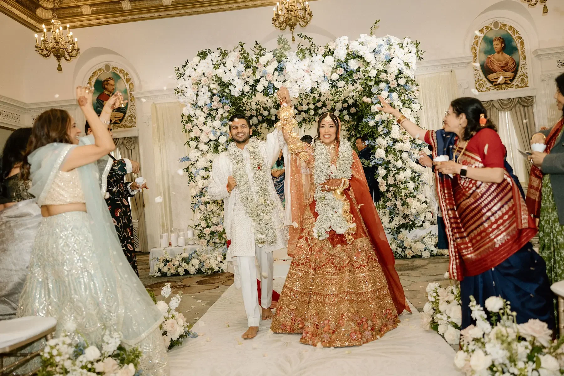 A bride and groom celebrating their wedding under a flower arch, with guests around them throwing flower petals.
