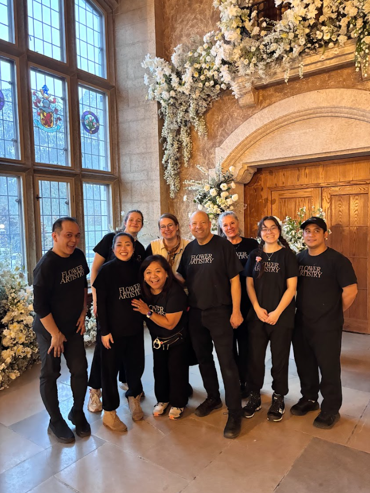 Group of people smiling inside a decorated venue with large stained glass windows and floral arrangements, possibly at a wedding or special event.