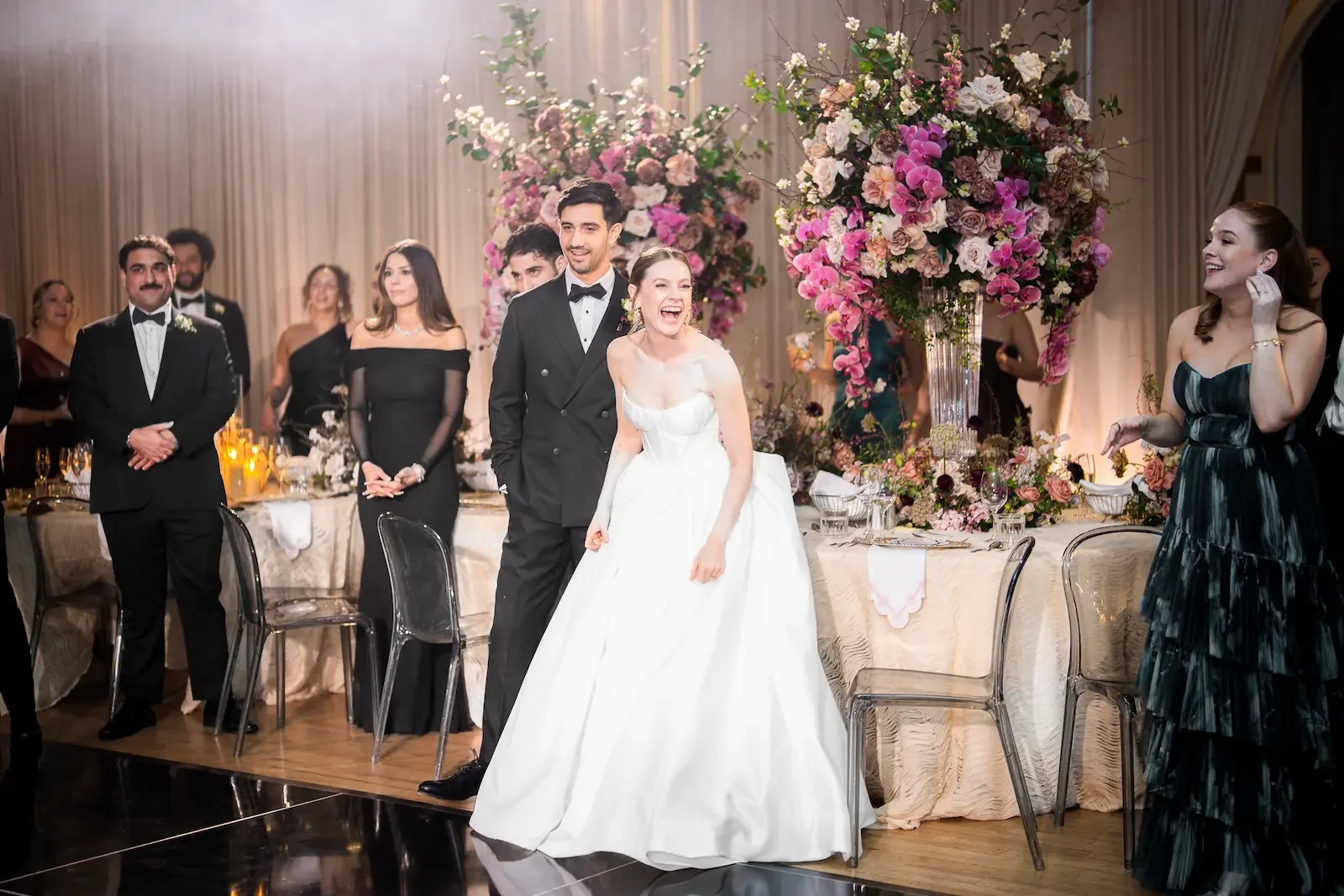 A wedding celebration with the bride and groom standing at a decorated banquet table, smiling and laughing. The bride is in a white wedding gown, and the groom is in a black tuxedo. Behind them are large floral arrangements with pink and white flowers, and guests are dressed in formal attire, enjoying the event.