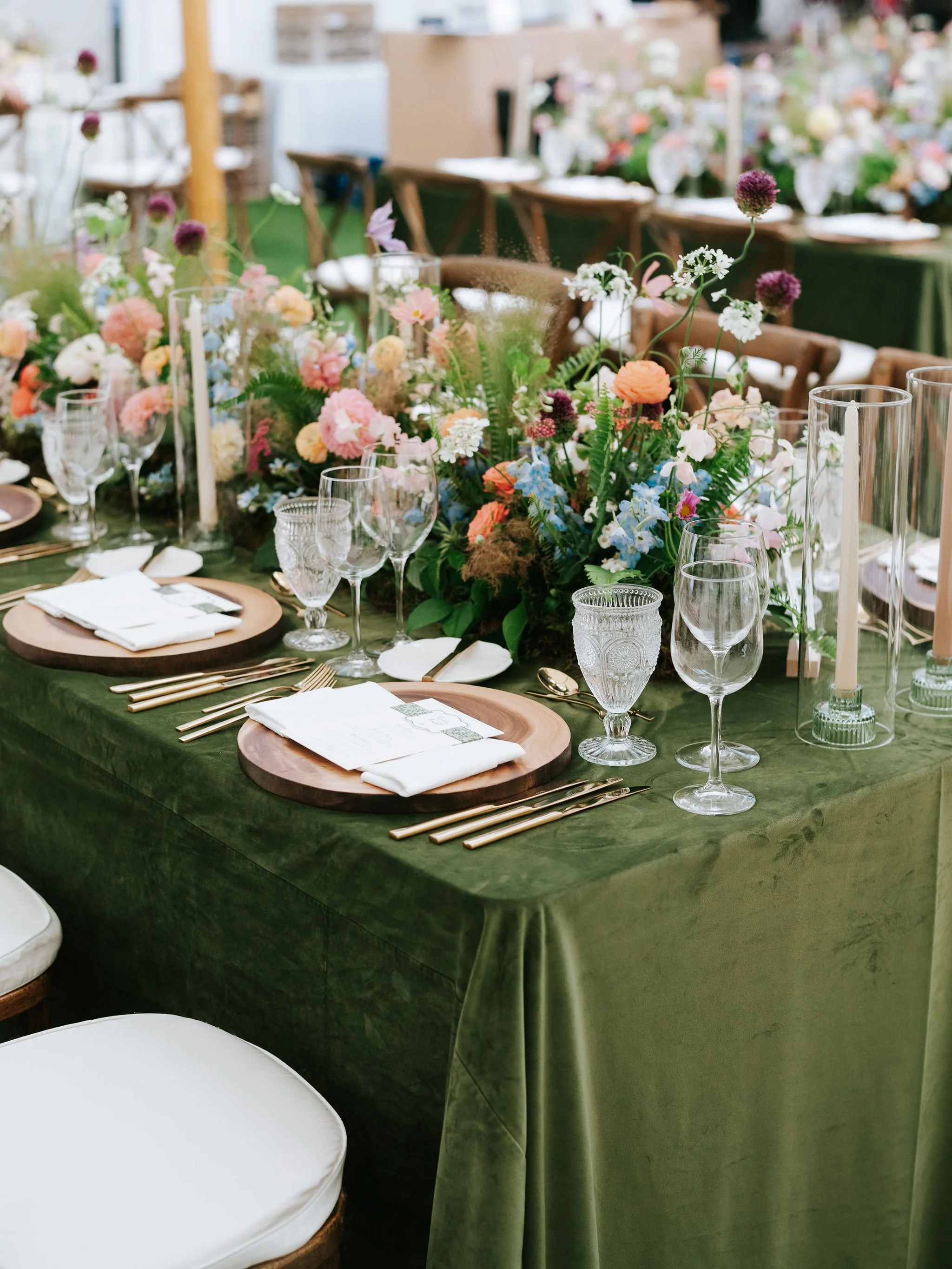 A table set for a formal event with a green tablecloth, floral centerpiece, crystal glasses, gold flatware, white napkins, and wooden chargers.