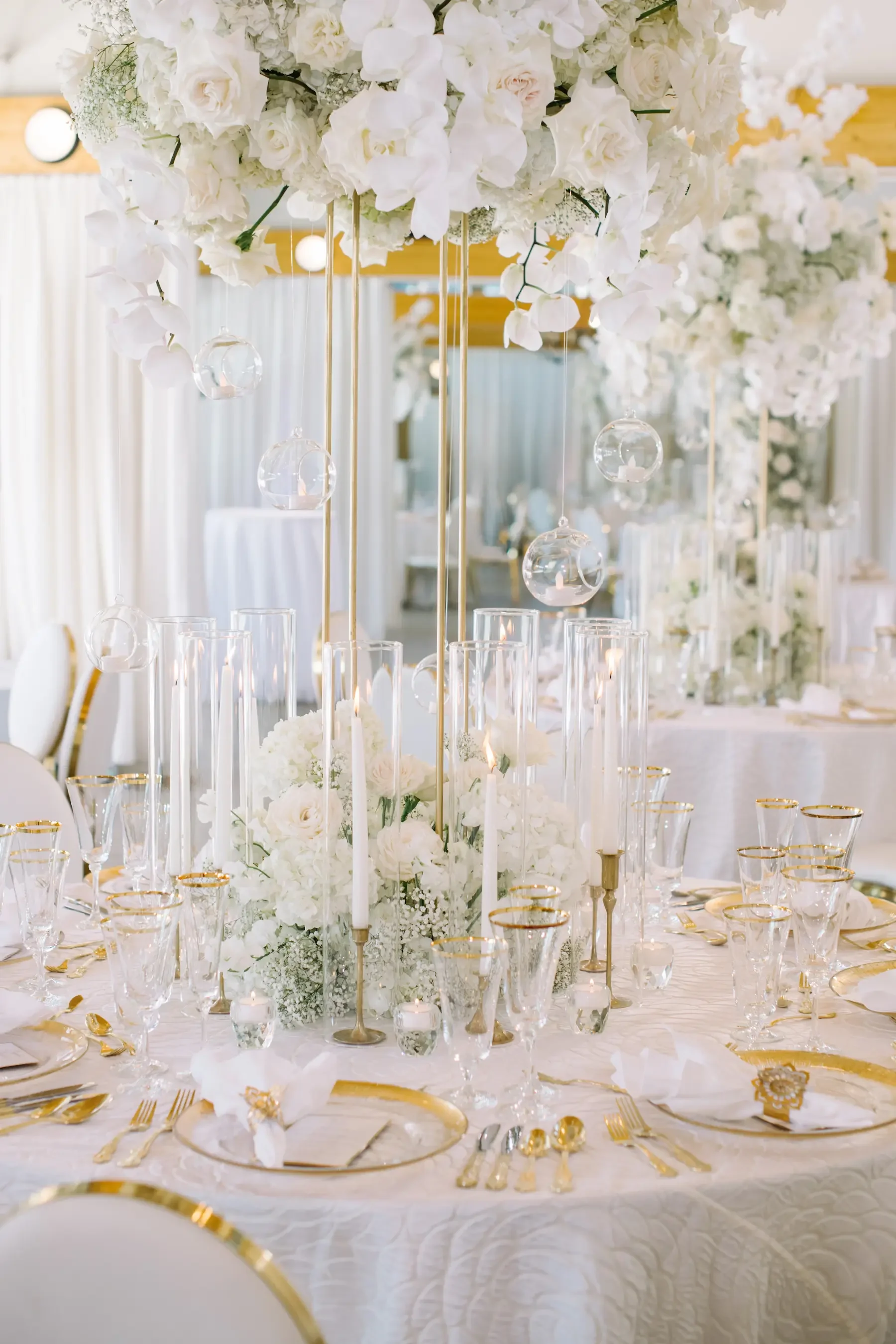 Elegant wedding reception table decorated with white flowers, glass candle holders, and hanging glass ornaments, with white and gold tableware.