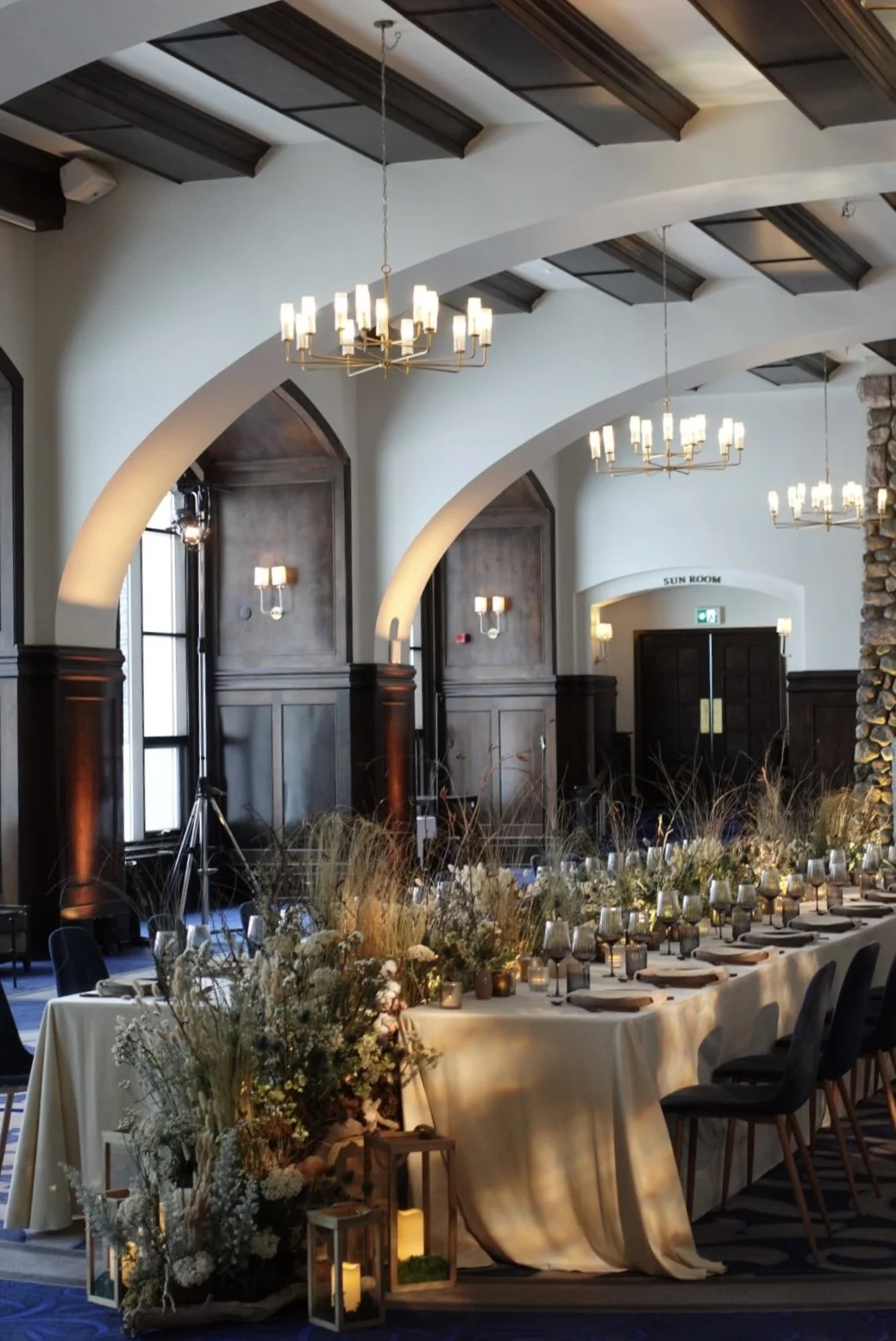 Elegant indoor event space with a long banquet table decorated with flowers, candles, and place settings, featuring high arched ceilings with dark wood beams and multiple chandeliers.