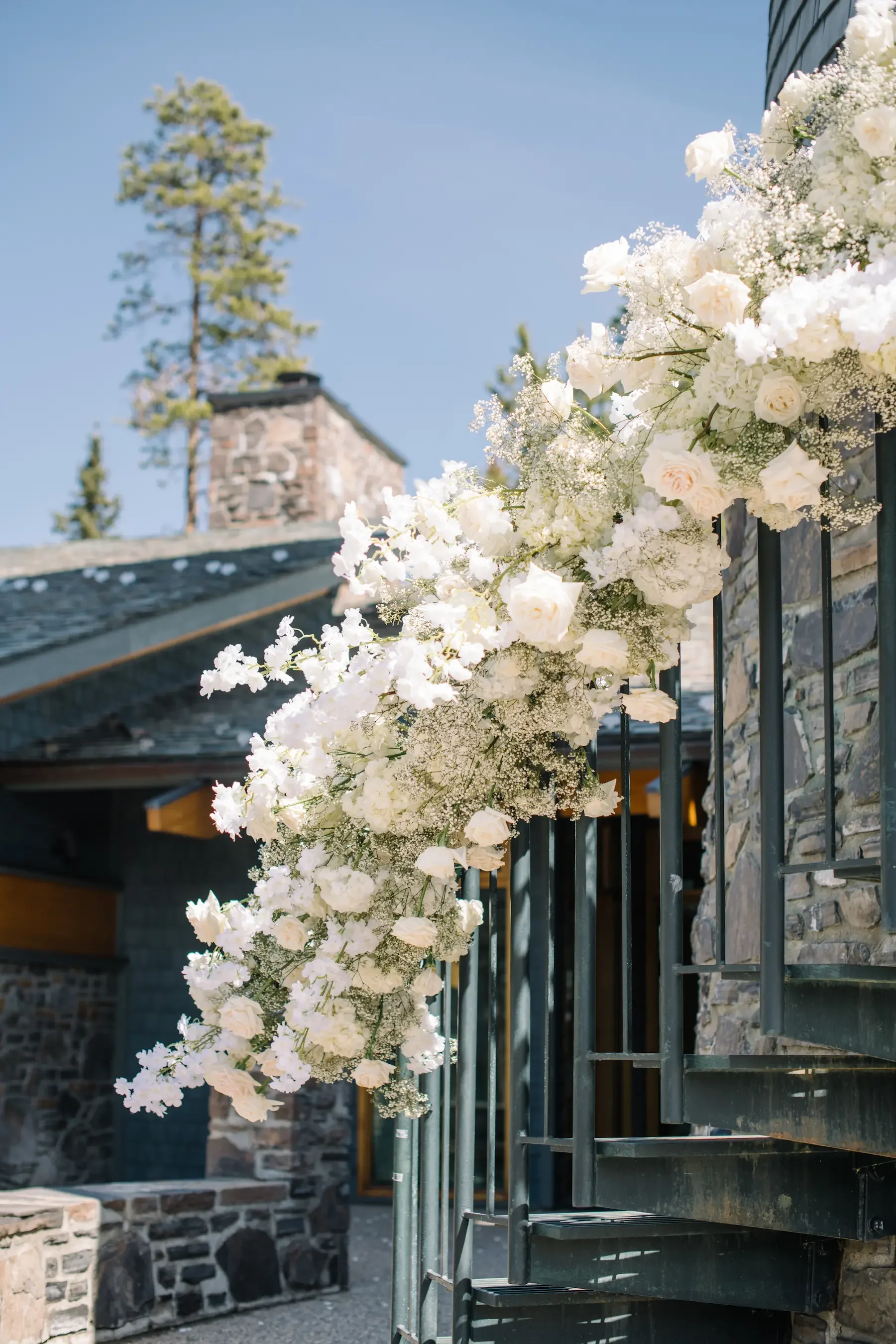 White floral arrangement on black metal stairs overlooking a stone building with a chimney, set against a clear blue sky.