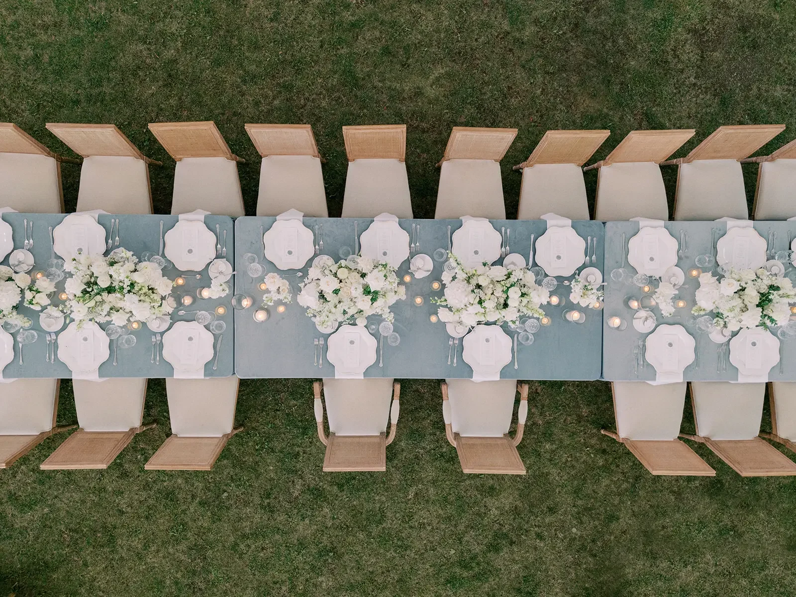 Overhead view of a long dining table with white floral centerpieces, candles, and white tableware, set on grass, surrounded by beige chairs with wooden backs.