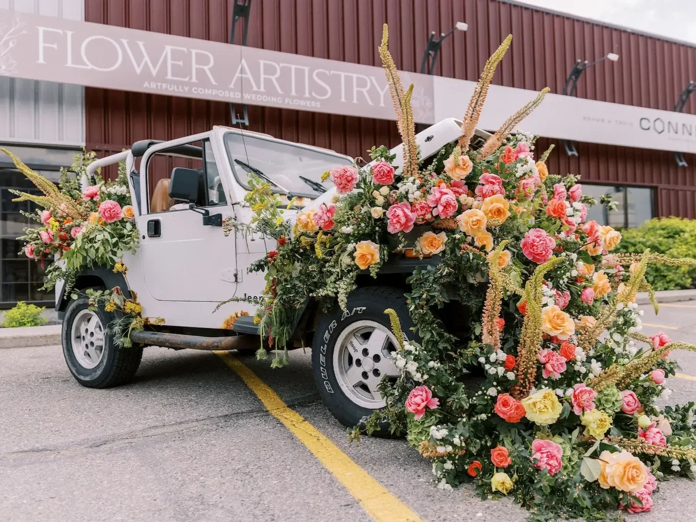 A white vintage jeep decorated with large, colorful flower arrangements, including pink, yellow, and orange roses and various greenery, parked in a lot in front of a flower artistry shop.