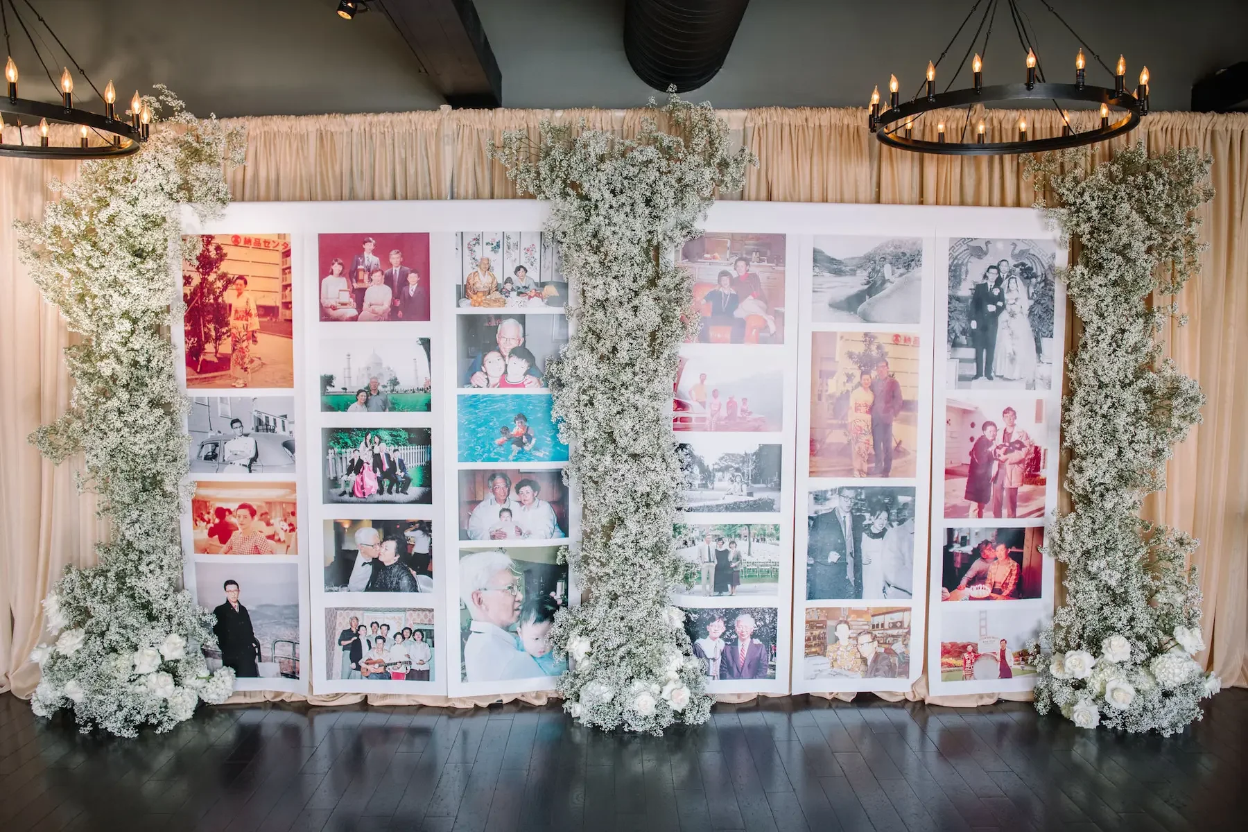 A collage of family photographs framed with white paper, decorated with white flowers arranged around the edges, set against a beige curtain backdrop at an event.