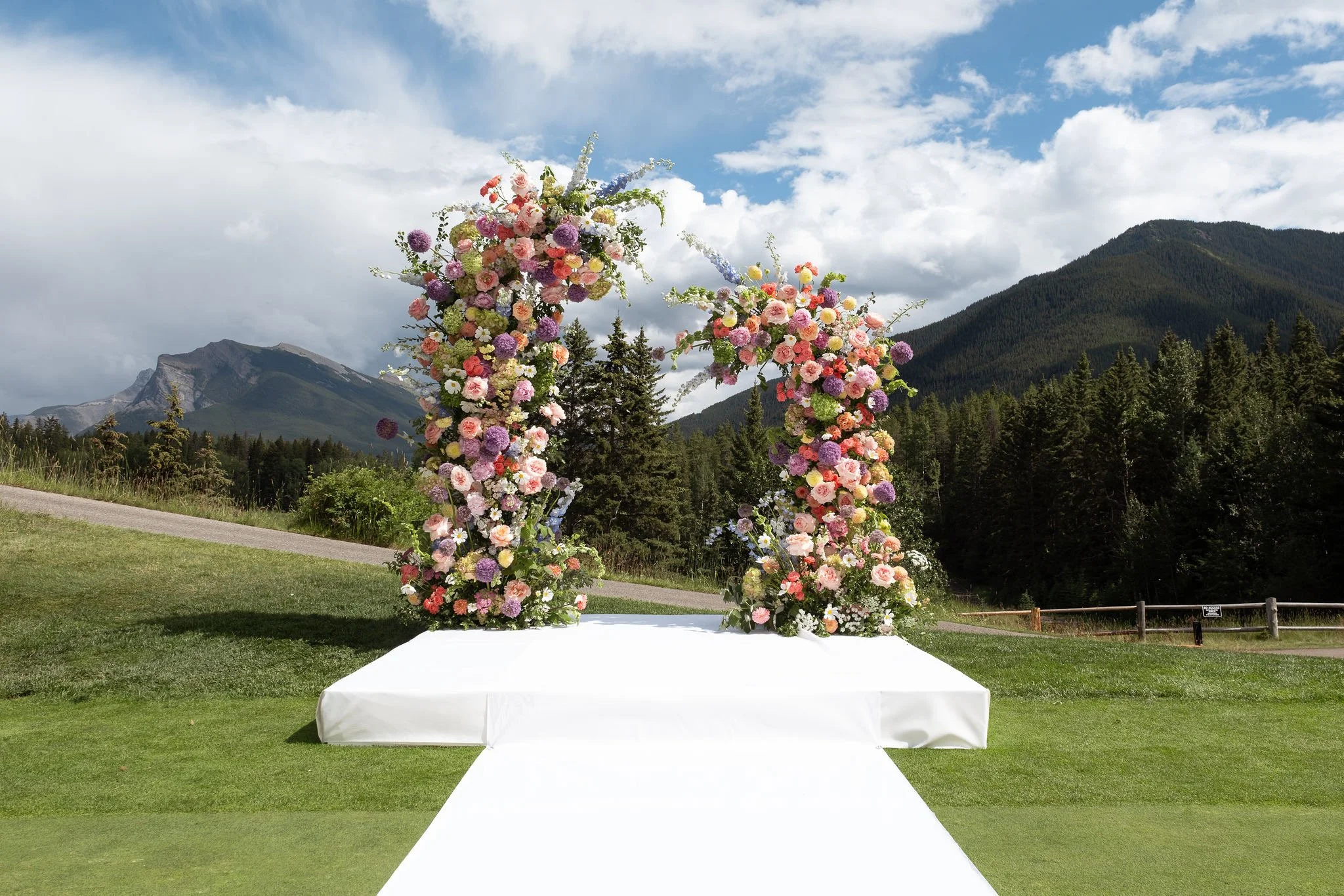 Outdoor wedding arch decorated with pink, purple, and white flowers on a grassy field, with mountains, trees, and a blue sky with clouds in the background.