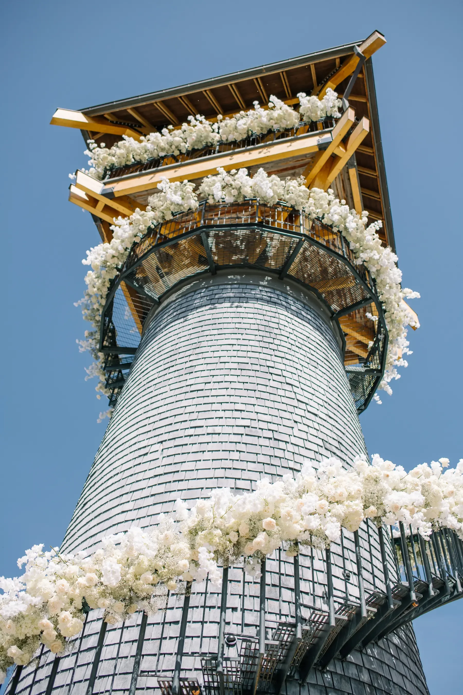 Close-up of a tall tower with gray shingles, white flowers decorating the railings, and wooden sections at the top, against a clear blue sky.