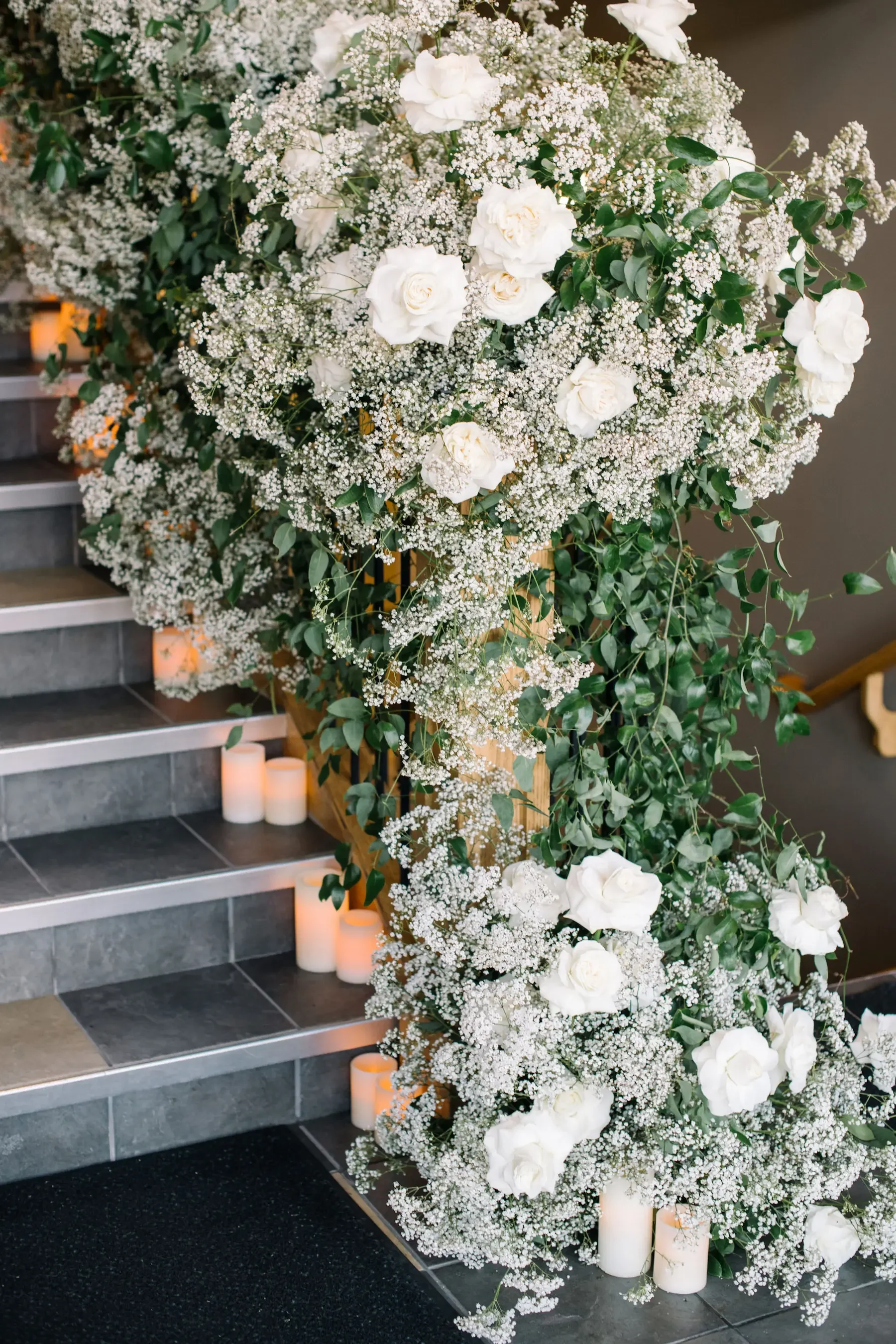Large floral arrangement with white roses, baby's breath, and greenery on stairs decorated with candles.