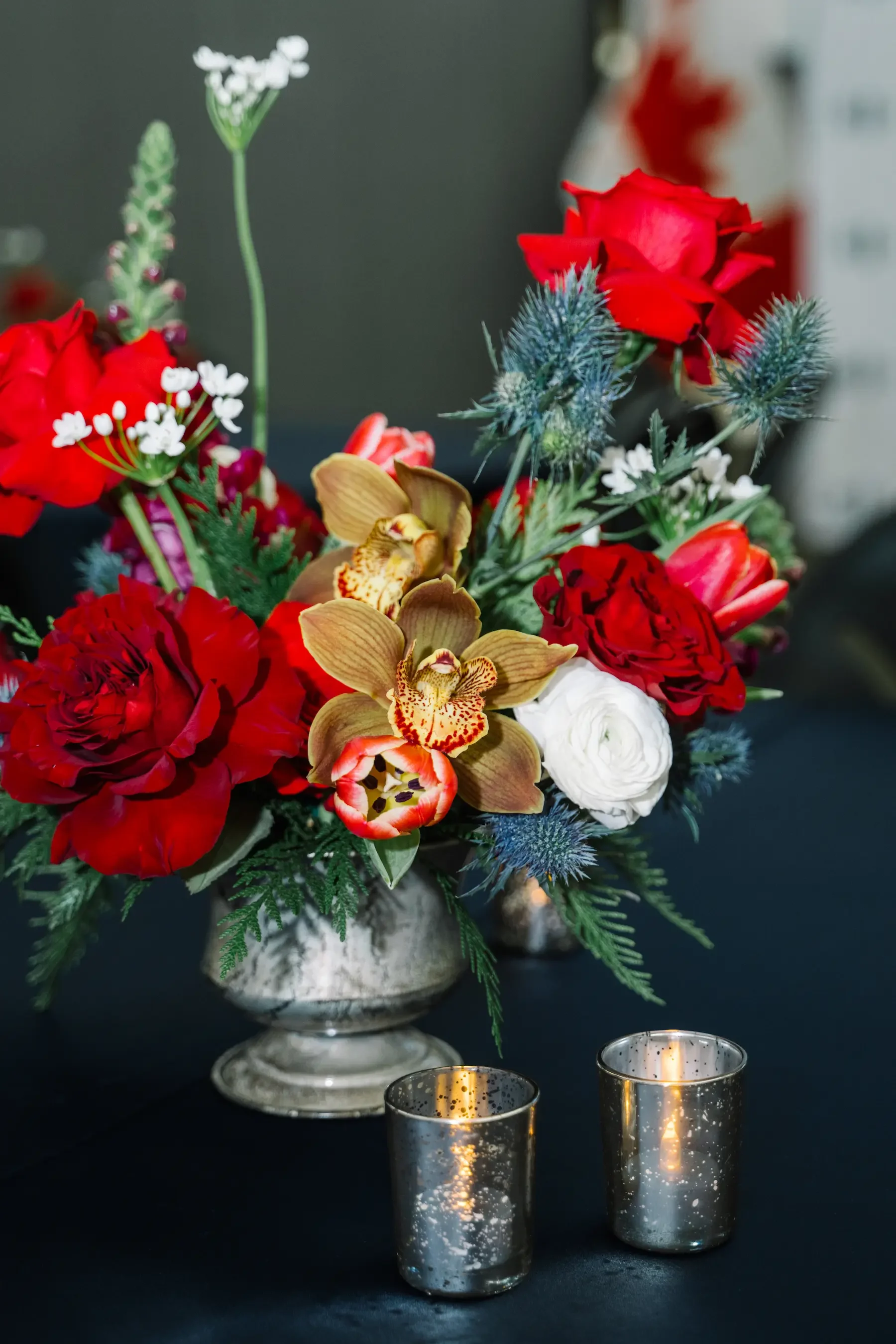 A floral arrangement with red, white, and tan flowers in a silver vase, placed on a black table with two small candle holders in front.