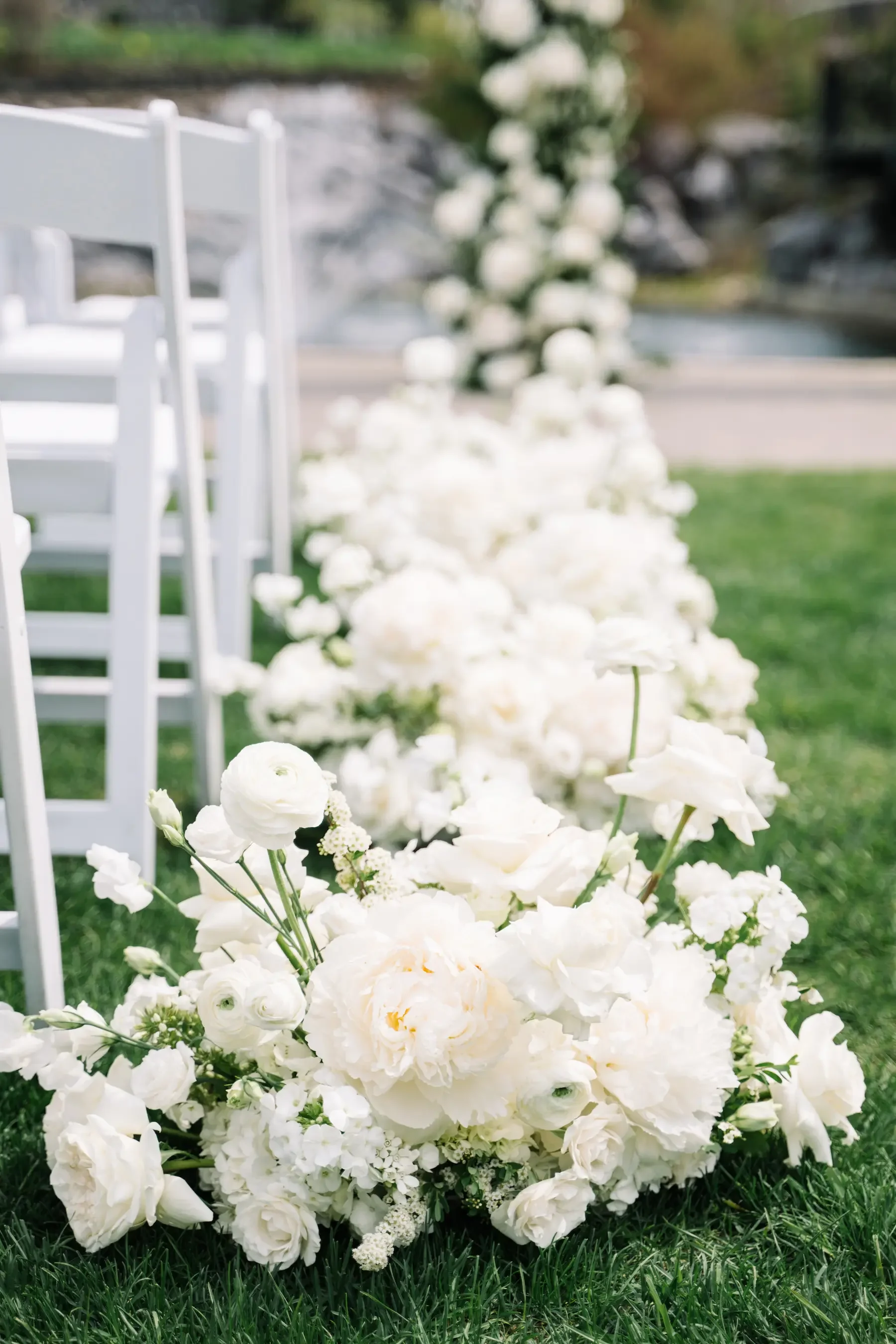 White floral arrangement on green grass at an outdoor wedding setting with white chairs in the background.