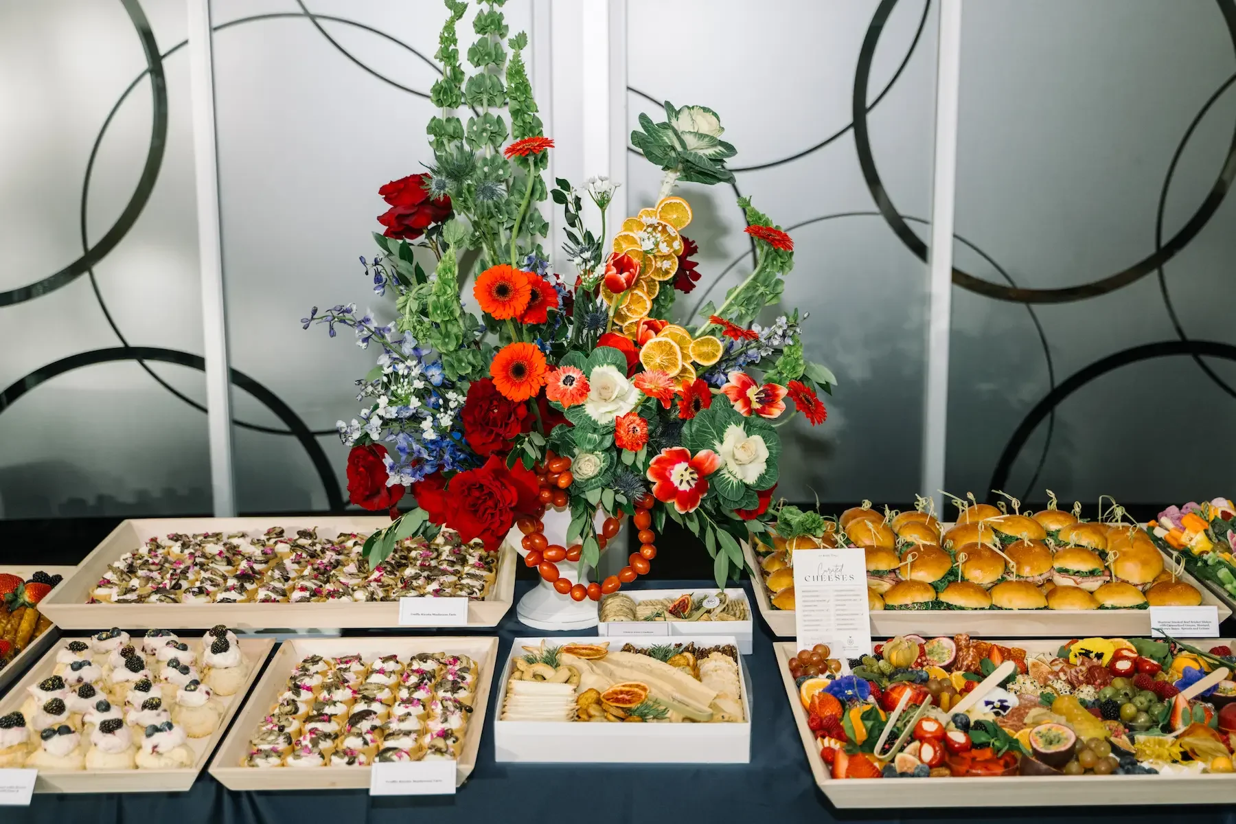 A buffet table with various desserts and finger foods, topped with a large colorful floral arrangement, on a black tablecloth with a modern geometric metal wall in the background.