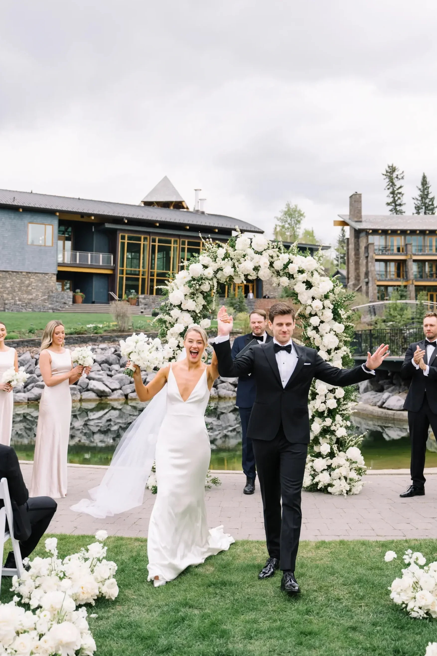 Bride and groom celebrating at their outdoor wedding ceremony, with bridesmaids and groomsmen, against a scenic backdrop with a floral arch and modern buildings.