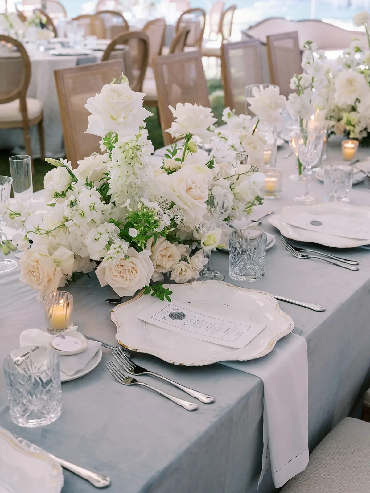Elegant wedding reception table with a large white floral centerpiece, candle votives, crystal glassware, and white plates with printed menus, set in a bright, outdoor venue with wooden chairs in the background.