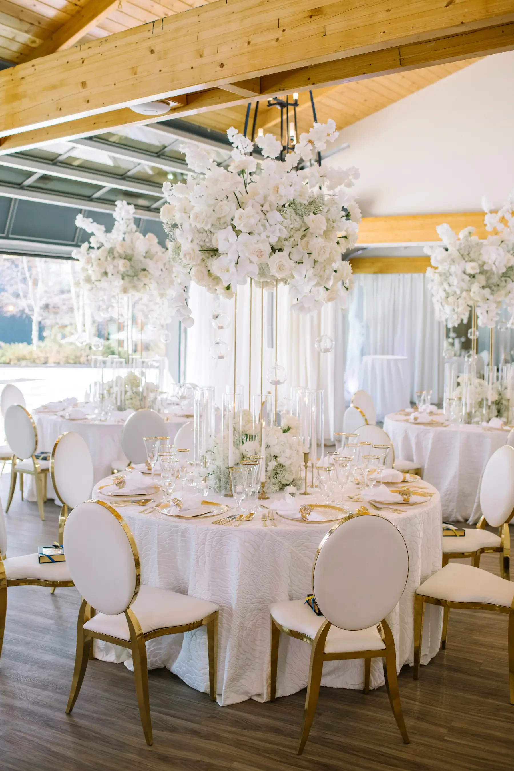 Elegant wedding reception tables with all-white floral centerpieces, gold-rimmed dinnerware, and white chairs with gold accents, inside a decorated event space with wooden beams and large windows.
