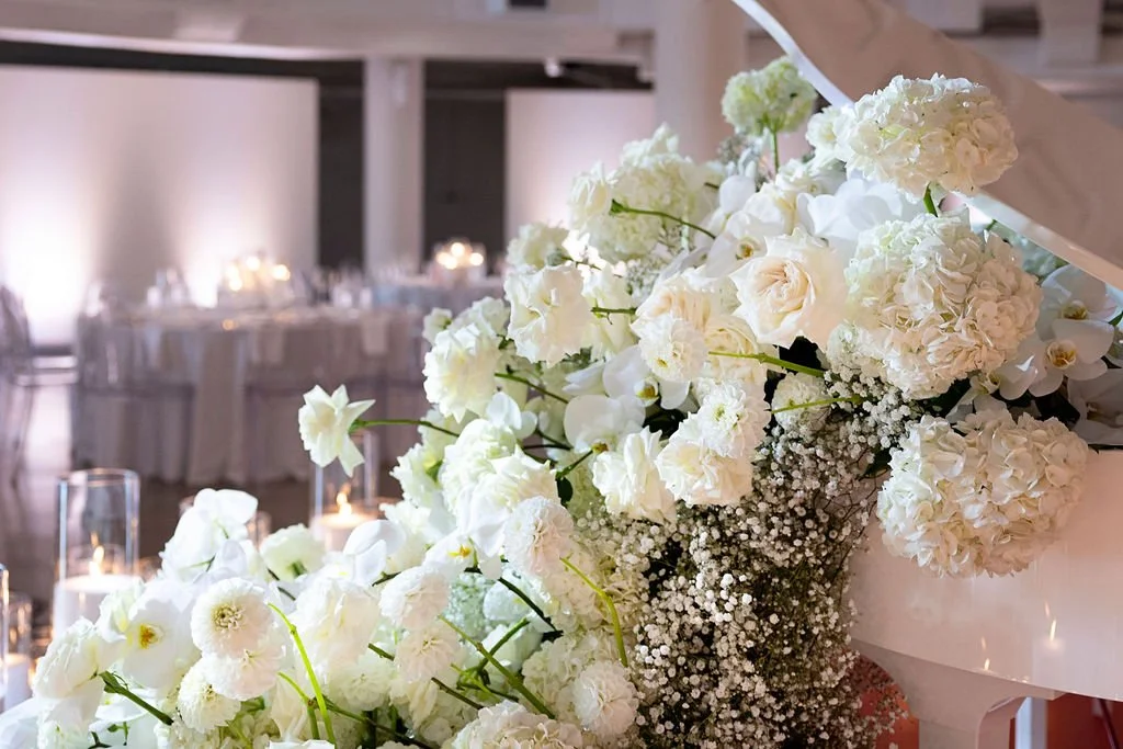 White floral arrangement on a piano at a wedding reception with decorated tables and candles in the background.