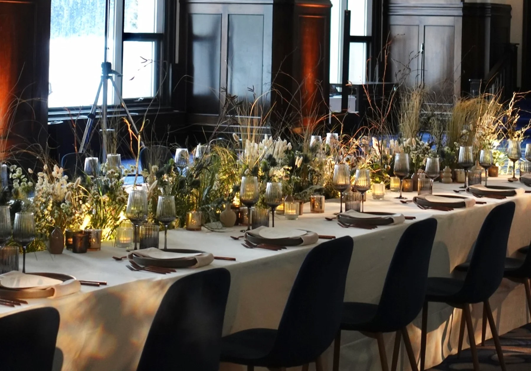 Long dining table set with white tablecloth, white plates, brown napkins, wine glasses, surrounded by black chairs, decorated with floral arrangements, candles, and lighting in a dark wood-paneled room.