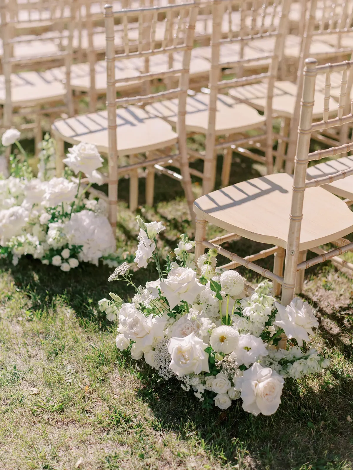 An outdoor wedding ceremony setup with beige chairs arranged in rows and white floral arrangements on the grass at the front.