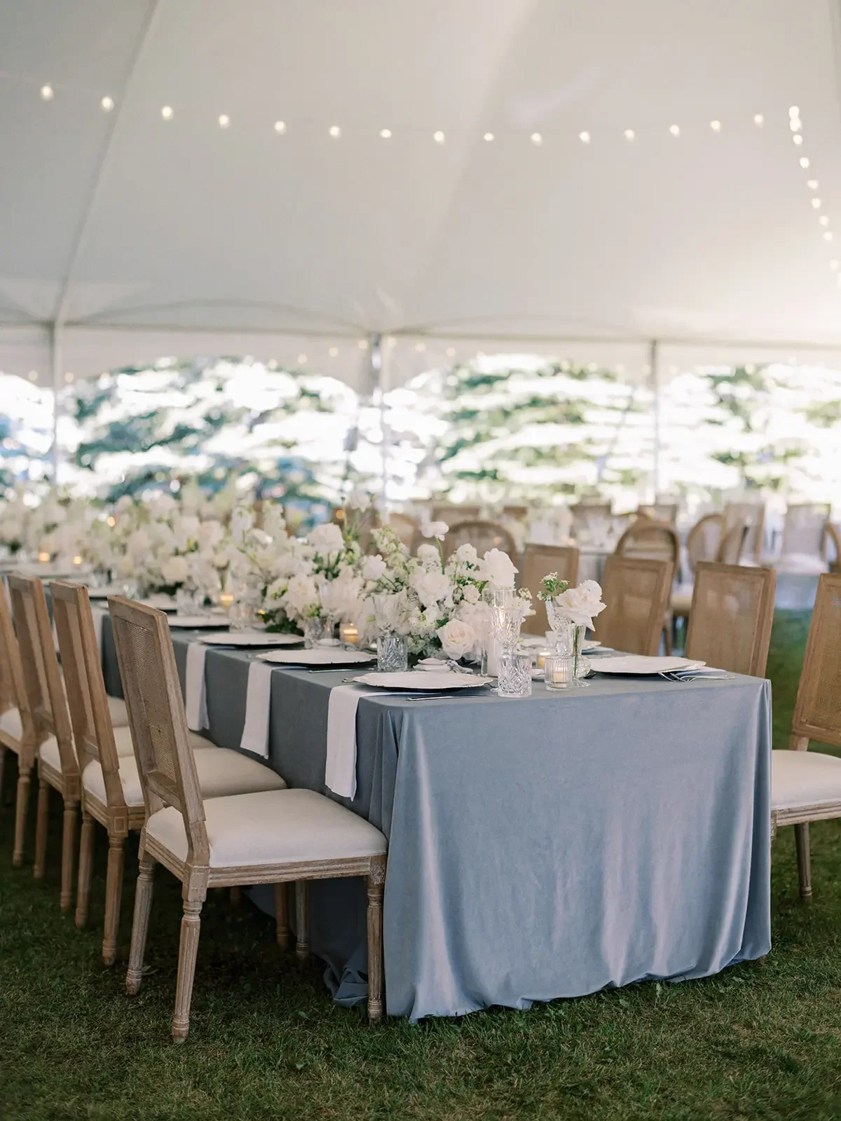 Elegant outdoor event setup with a long table covered in a gray tablecloth, decorated with white floral arrangements and candles, under a white tent with string lights.