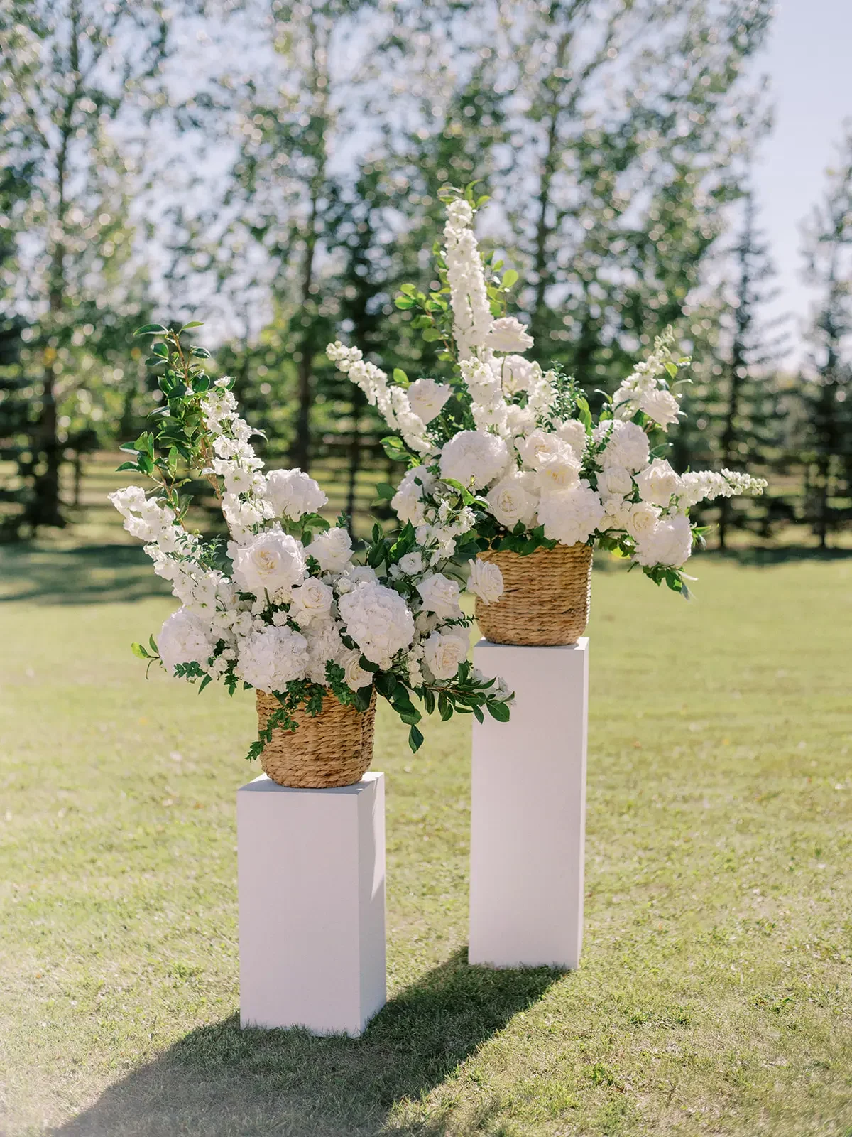Two floral arrangements with white flowers in woven baskets placed on white pedestals outdoors with trees in the background.