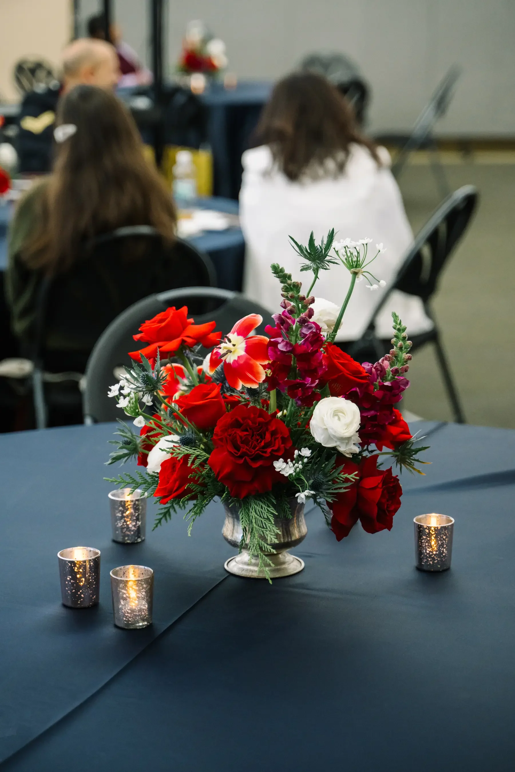 A floral centerpiece with red, white, and purple flowers on a dark table, surrounded by four small lit candles, at an indoor event with blurred attendees in the background.