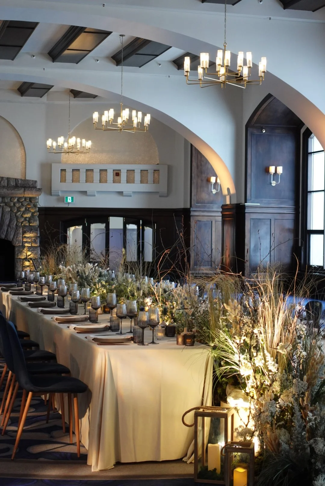 Elegant indoor event space with a long dining table, set with black chairs, glassware, plates, and napkins. The table is decorated with floral arrangements and candles. The room features high ceilings with chandeliers, arched windows, and dark wooden wall paneling.