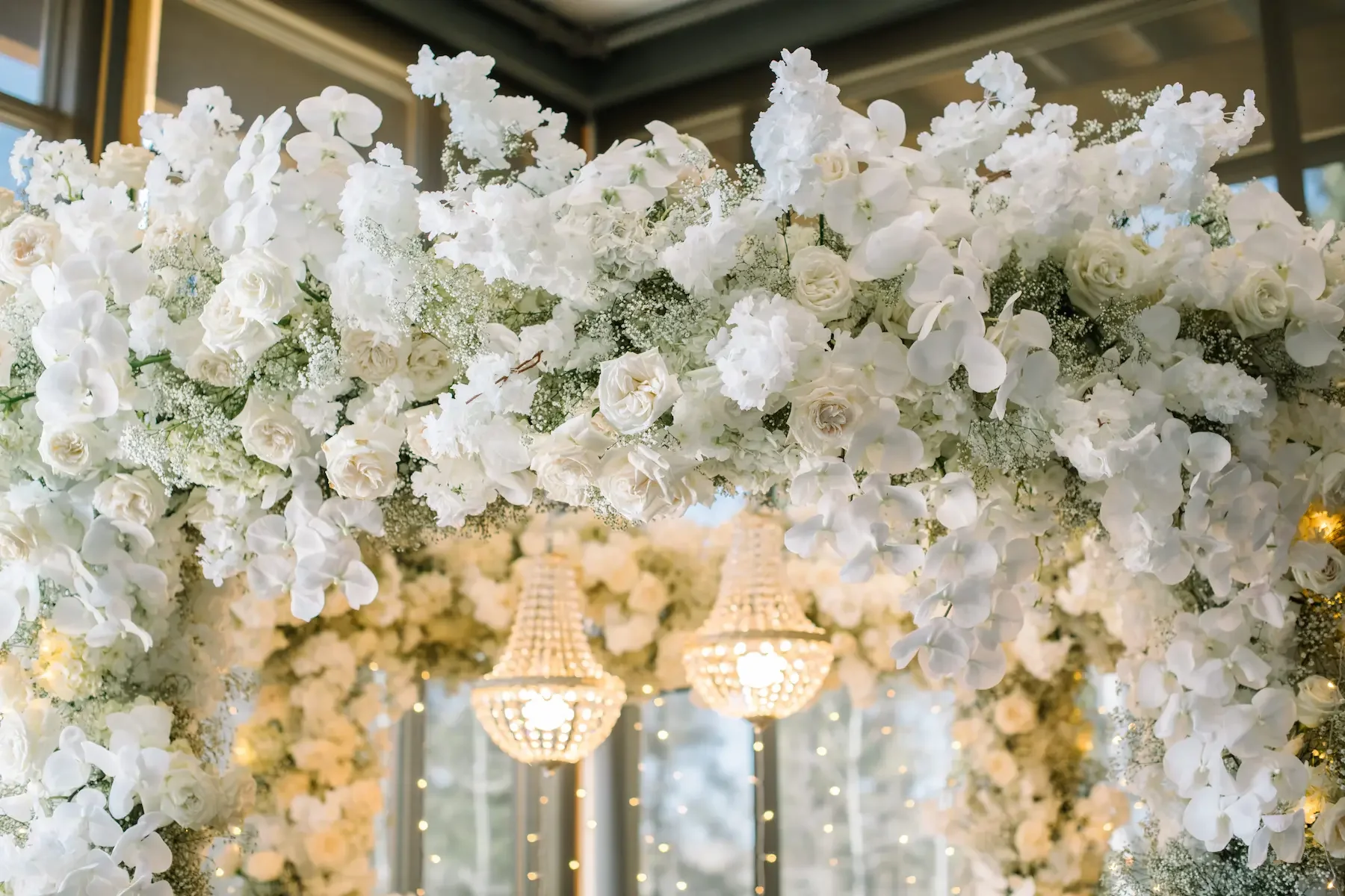 White floral arch with white roses, orchids, and baby's breath, decorated with chandeliers and string lights in a room with large windows.