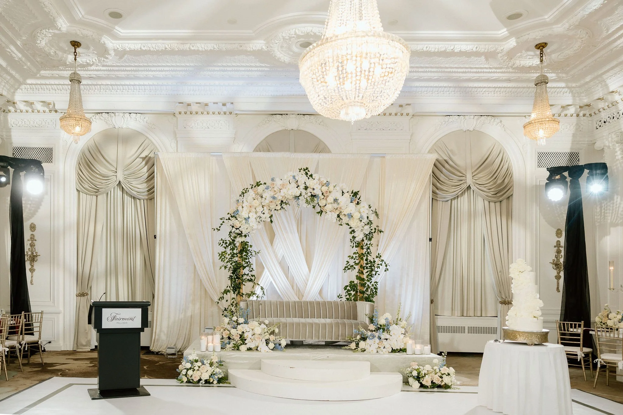 Elegant wedding ceremony setup with a floral arch, draped curtains, chandelier, and decorated stage with a white cake and candles.