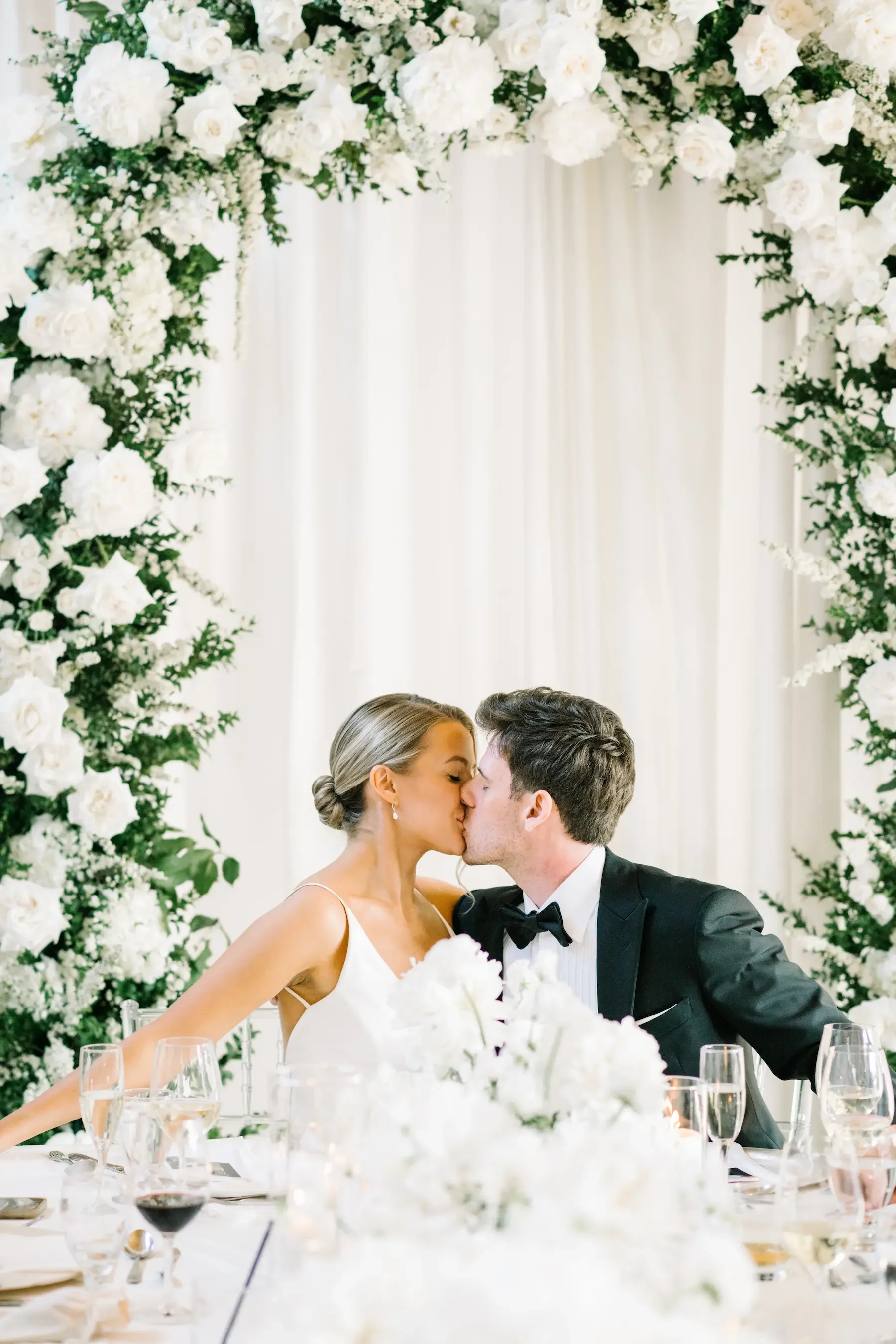 A bride and groom sharing a kiss at their wedding reception, surrounded by white floral decorations and elegant table settings.