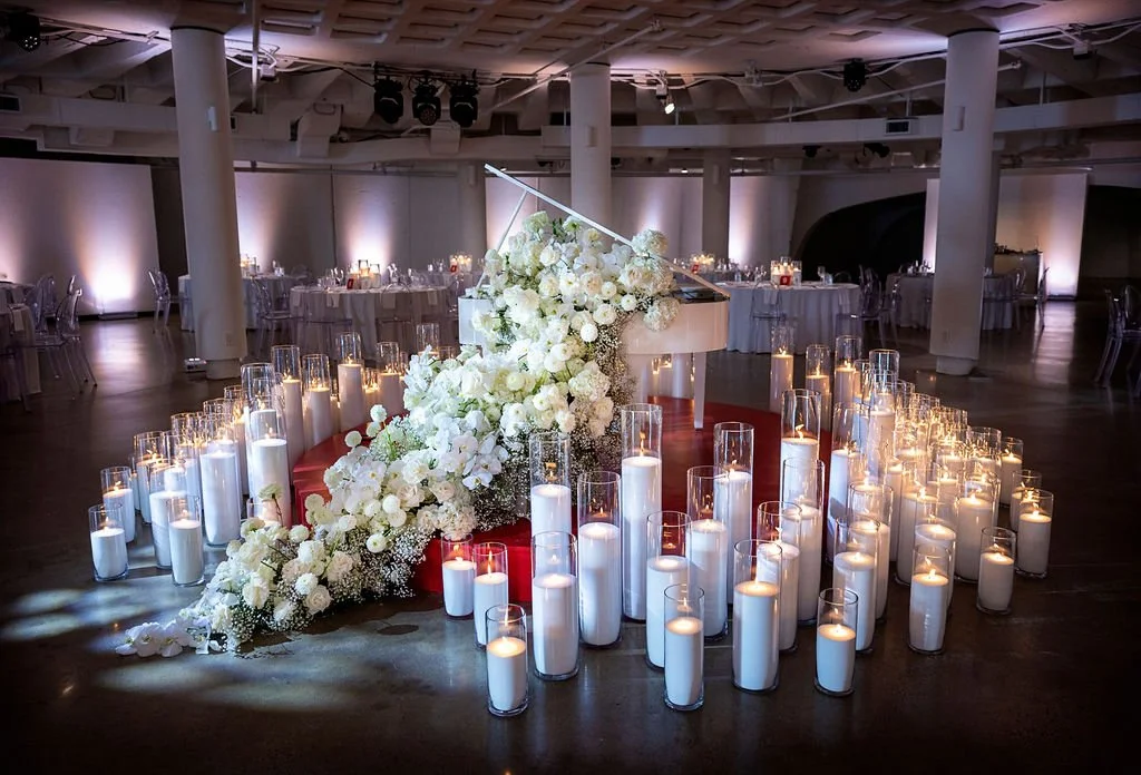 A grand piano decorated with a cascading white floral arrangement surrounded by numerous white candles in glass holders, set in a large, elegant banquet hall with dim lighting and round tables.