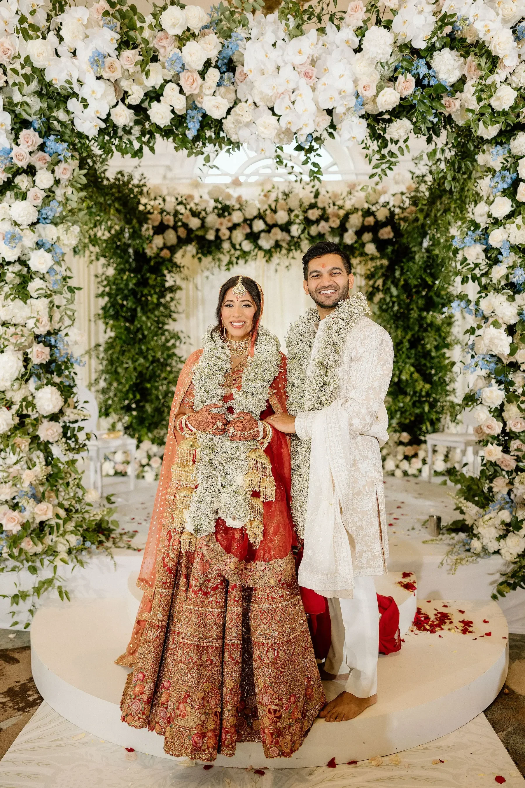 A bride and groom in traditional Indian wedding attire standing beneath a floral arch with white, pink, and blue flowers, smiling for a photo.