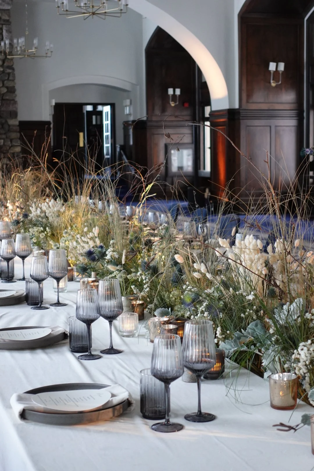 Elegant dining table set with gray glasses, plates, candles, and a lush floral centerpiece in a decorated room with dark wood paneling and white walls.