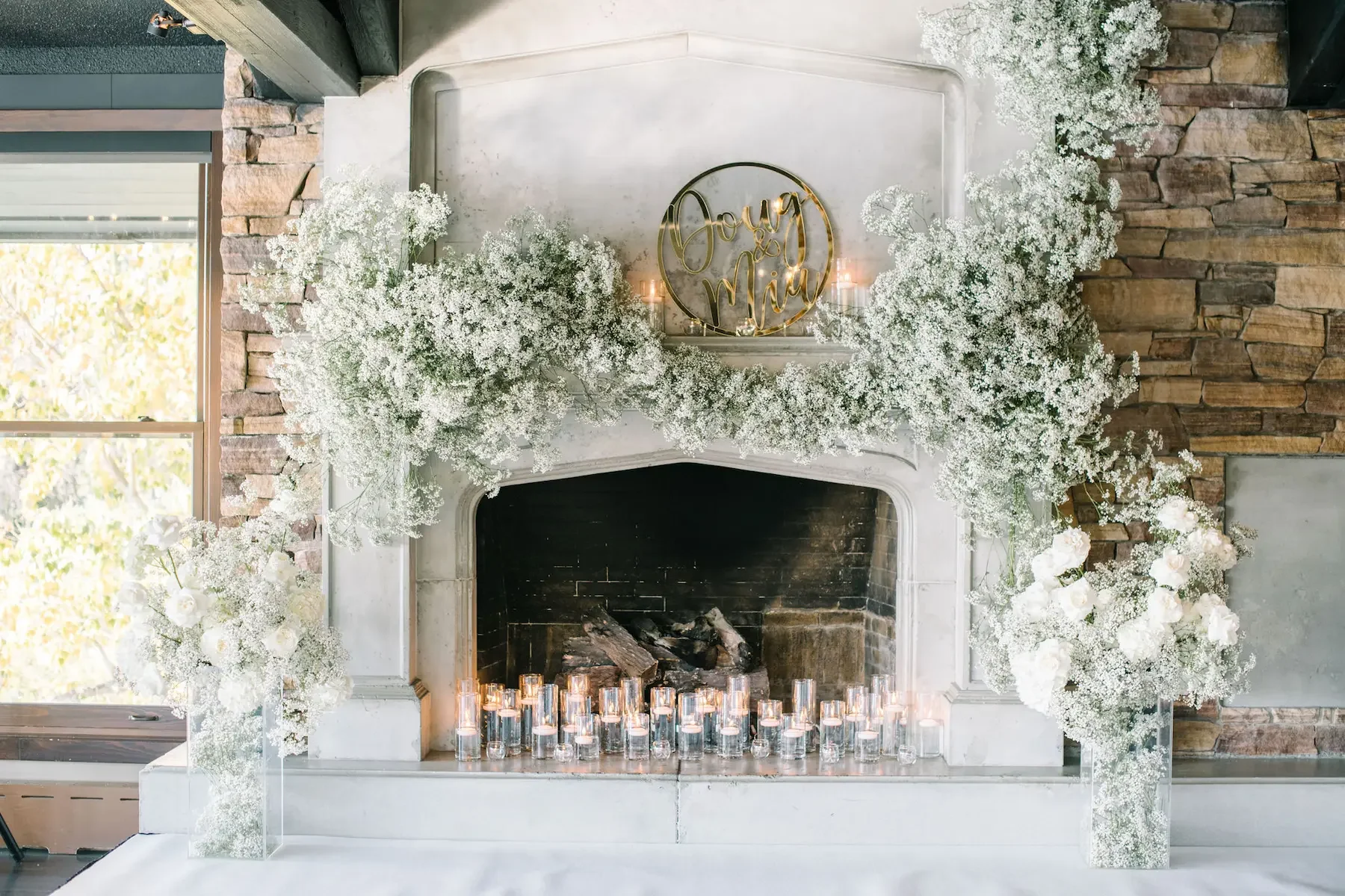 Decorated fireplace with white floral arrangements and candles, wedding sign reading 'Boy & Mia' on the mantle, window with blinds to the left, and brick wall to the right.