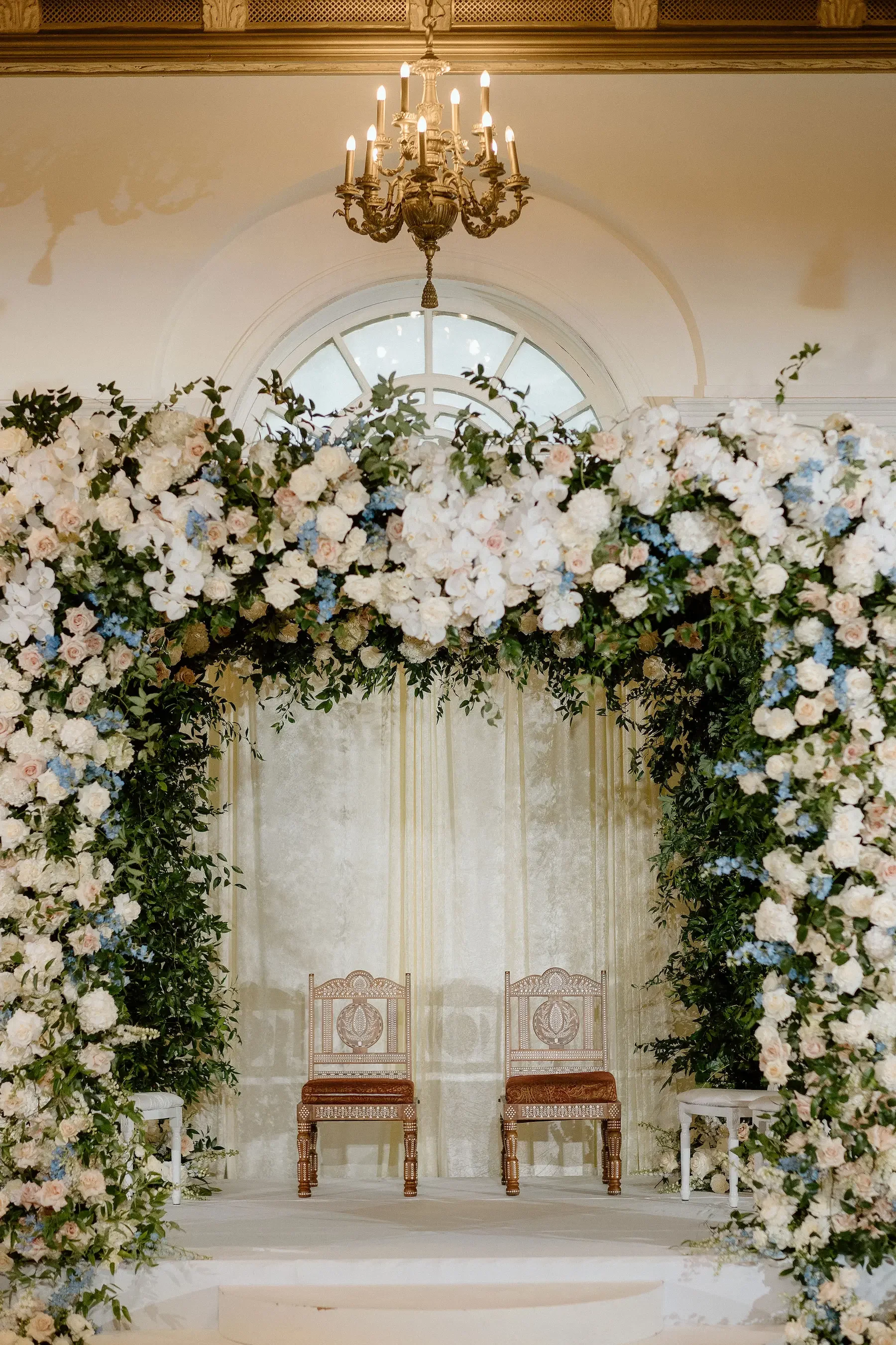 Wedding altar decorated with a large floral arch of white and pale pink flowers and green foliage, with two ornate wooden chairs on a stage, under a chandelier and beside a window.