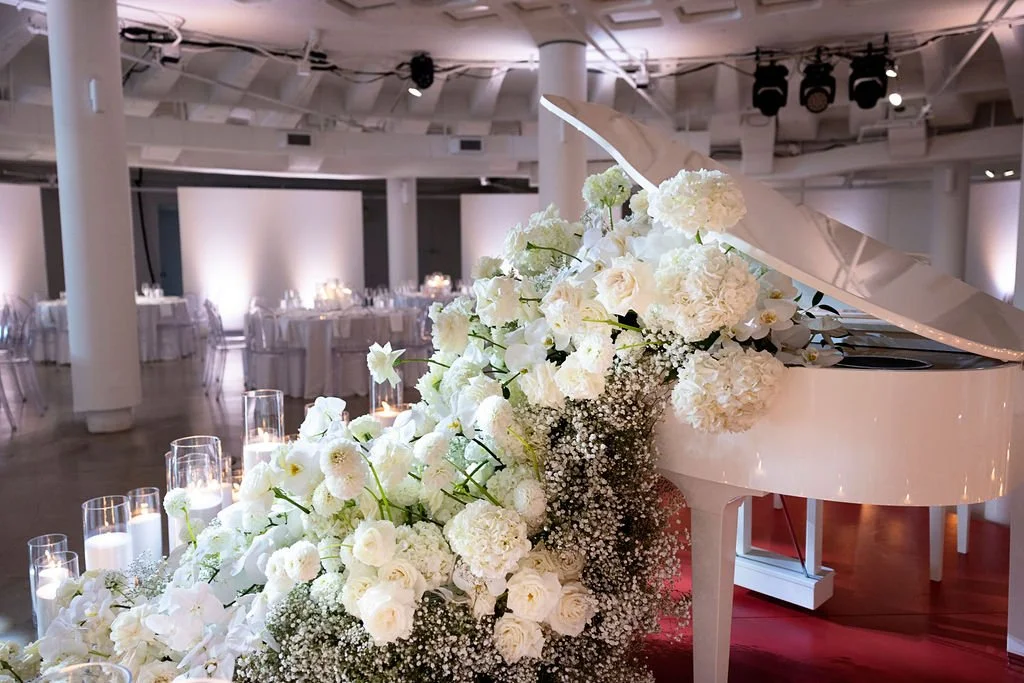 White grand piano decorated with white flowers, including roses and hydrangeas, in a banquet hall set for an event.