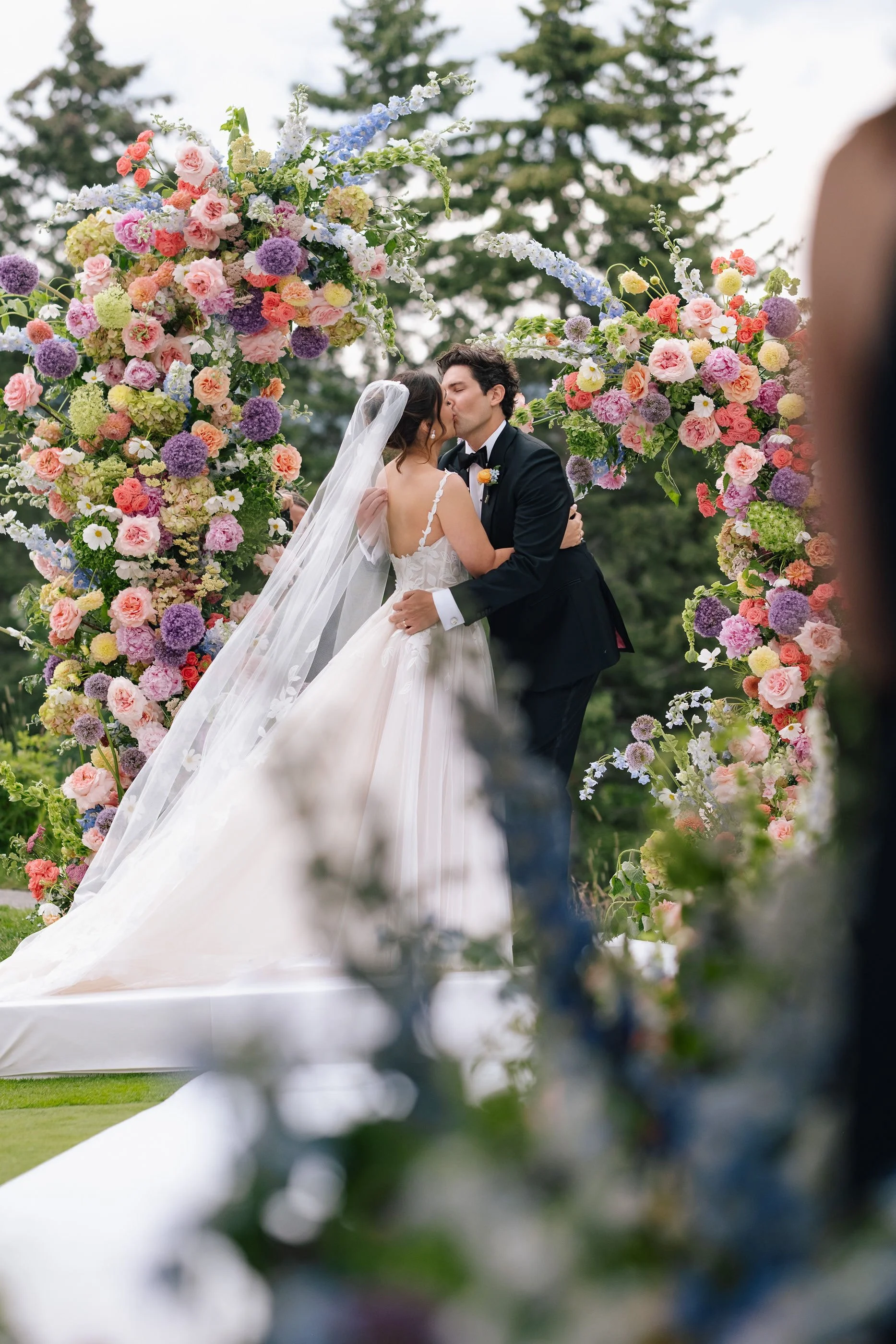 A bride and groom sharing a kiss during their outdoor wedding ceremony, surrounded by a colorful floral arch with pink, purple, yellow, and white flowers, on a green grassy area with trees in the background.