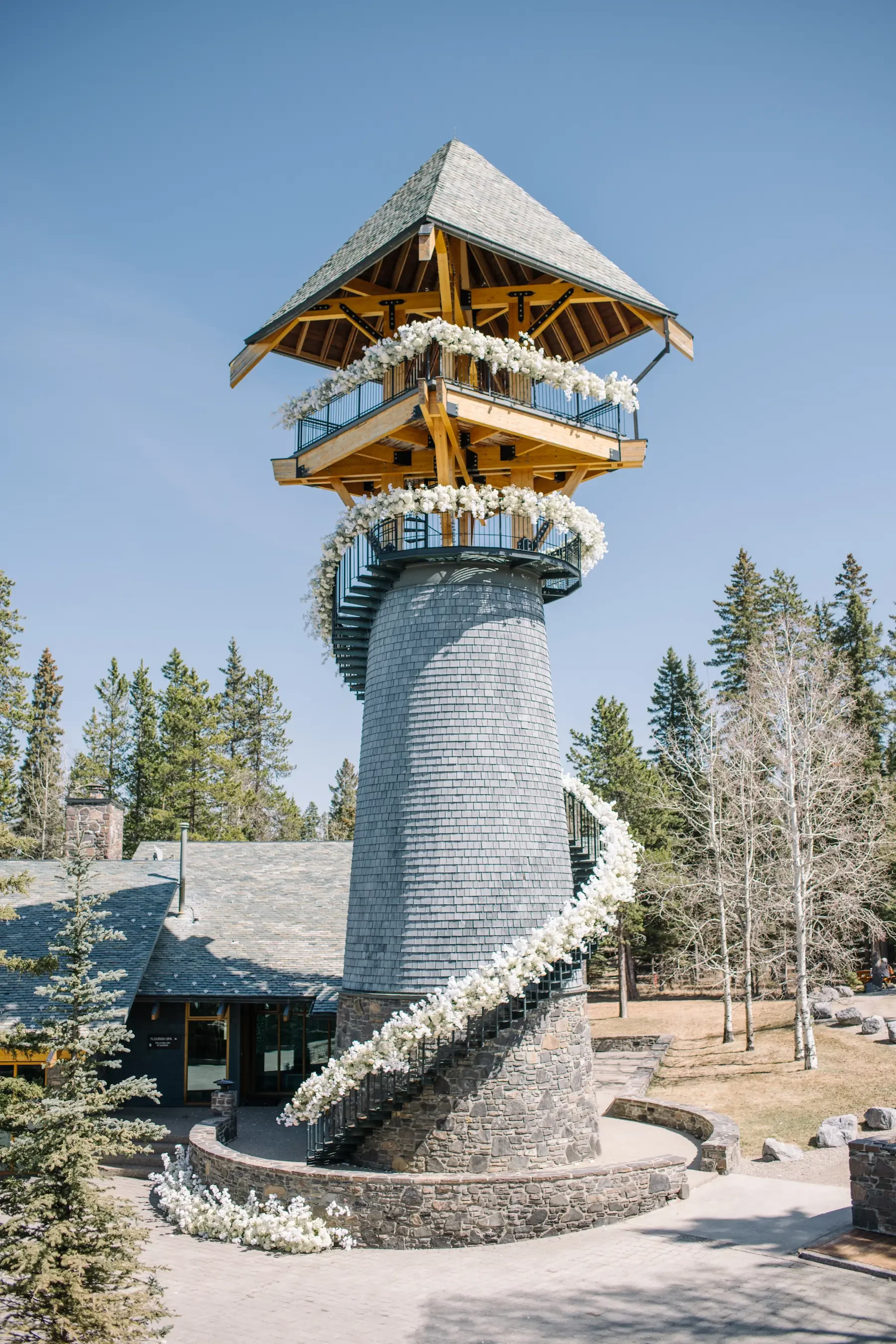 A tall, whimsical lighthouse-like tower with a spiral staircase wrapping around it, decorated with white flowers and greenery, set in a natural landscape with trees.