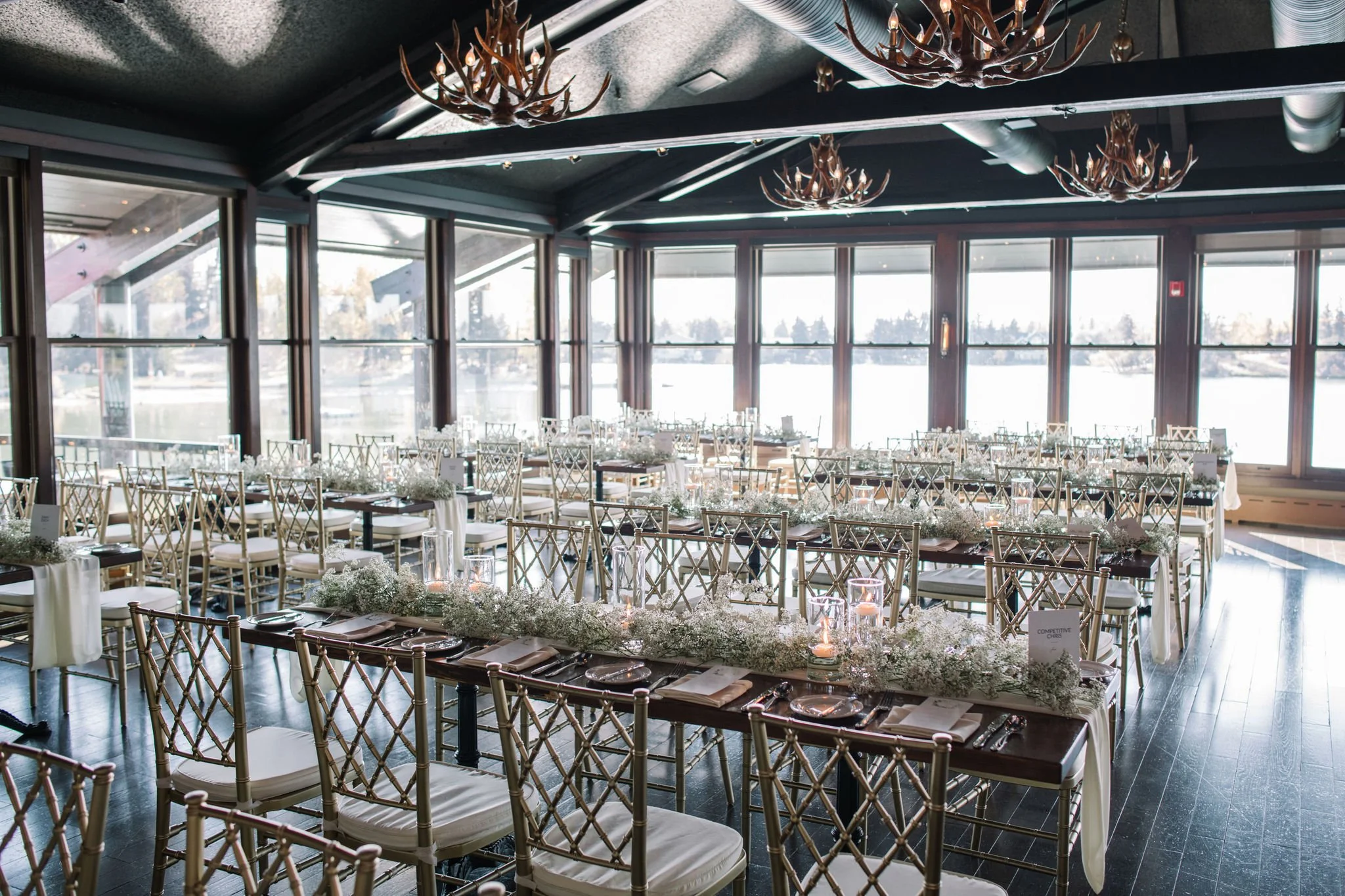 Interior of a banquet hall decorated for an event with round and rectangular tables, gold lattice-back chairs, floral centerpieces, candles, and large windows overlooking a snowy outdoor scene with trees.