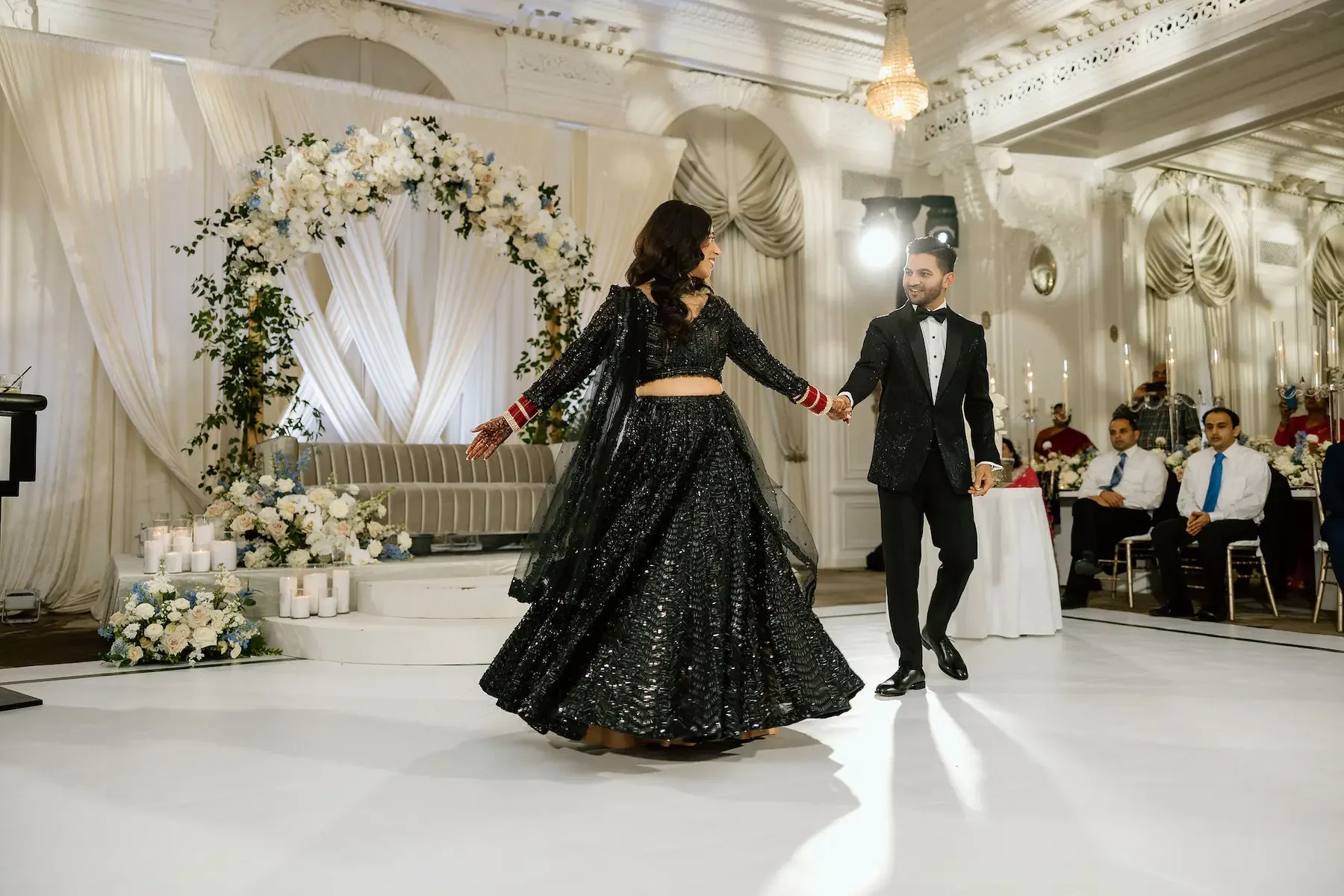A couple dancing at a wedding reception. The woman is wearing a black sequined traditional outfit and the man is in a black tuxedo. The background features elegant white drapes, floral arrangements, candles, and seated guests watching the dance.