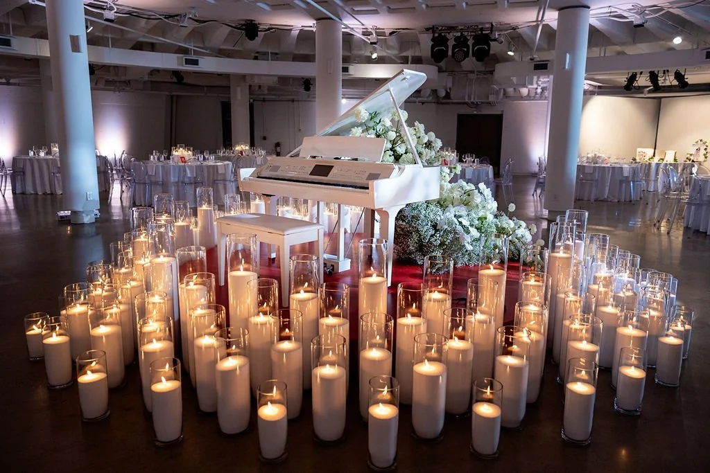 A white grand piano decorated with white flowers in the center of a large event hall. The piano is surrounded by numerous white candles in glass holders, creating a warm glow. The hall has tables with white tablecloths and chairs arranged in the background, suggesting a formal event or celebration.