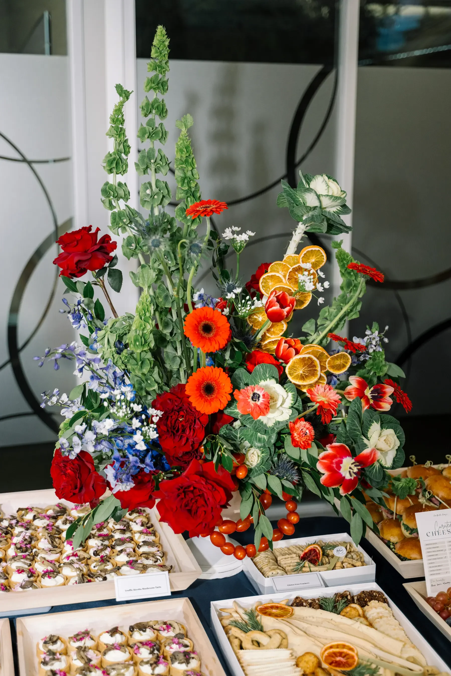 A large, colorful floral arrangement on a table with various desserts, including cookies and cheeses, against a modern background.