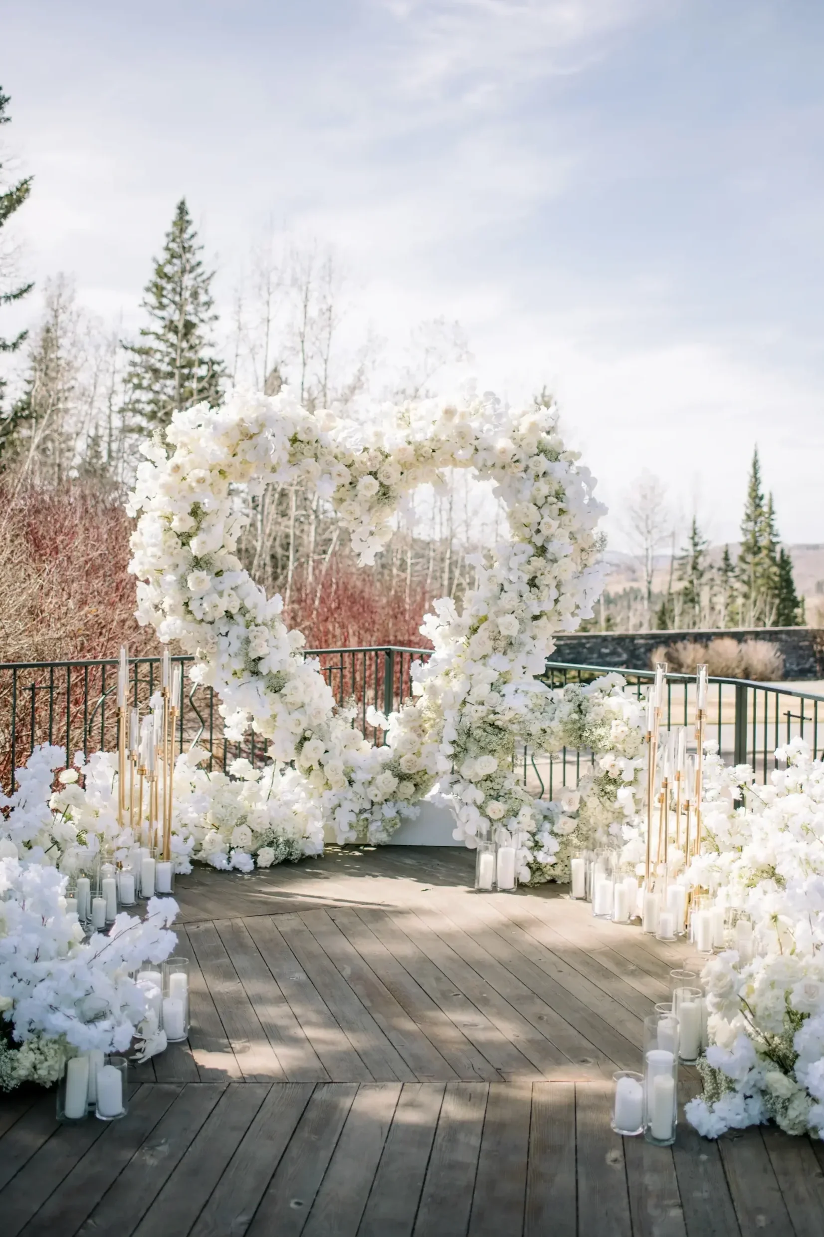 White floral heart-shaped wedding arch surrounded by white flowers and candles on a wooden platform outdoors.