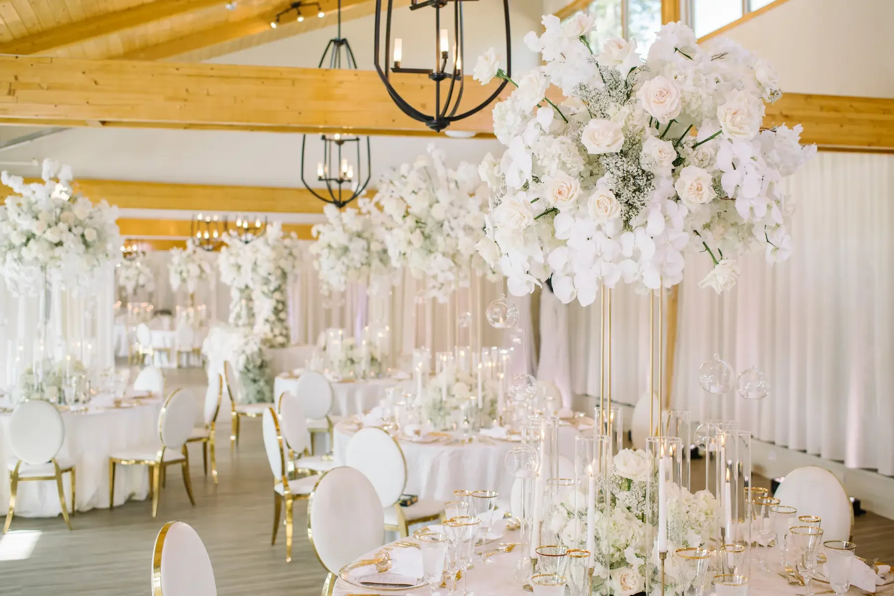 Elegant wedding reception decorated with tall floral arrangements of white roses and orchids, gold-rimmed glassware, and white table linens in a bright venue with wooden beams and chandelier lighting.