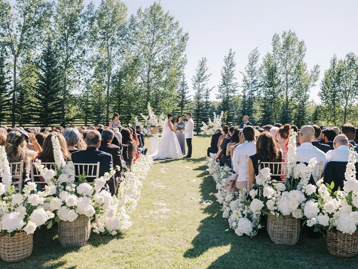 Outdoor wedding ceremony with a bride and groom standing at the altar, surrounded by seated guests, decorated with white flowers in baskets, set in a lush green garden under a clear blue sky.