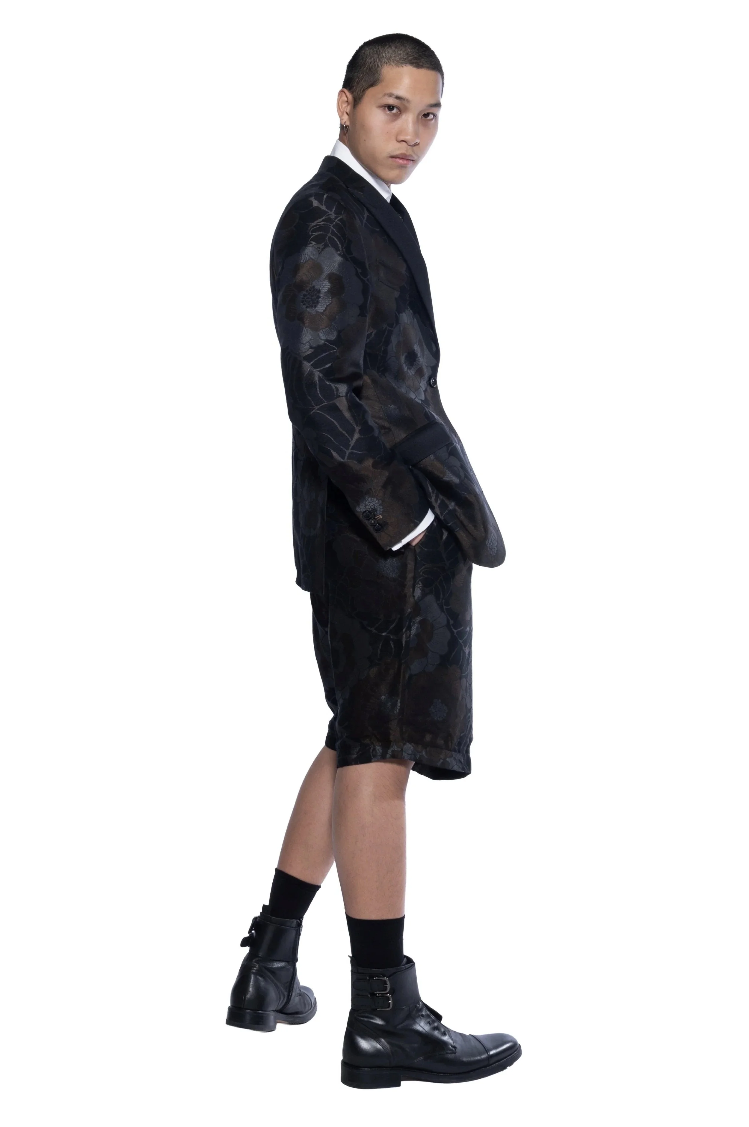 Un jeune homme porte un costume sombre avec un motif floral, des bottines en cuir et des chaussettes noires, debout contre un fond blanc, regardant la caméra avec une expression sérieuse.