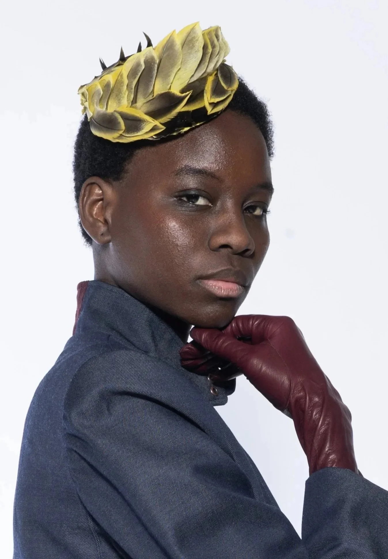 Portrait d'une femme noire portant une veste bleu denim, un gant en cuir bordeaux sur la main droite, et un accessoire en forme de chapeau en plume d'oiseaux jaune et noir sur la tête, posant avec la main sous le menton contre un fond blanc.