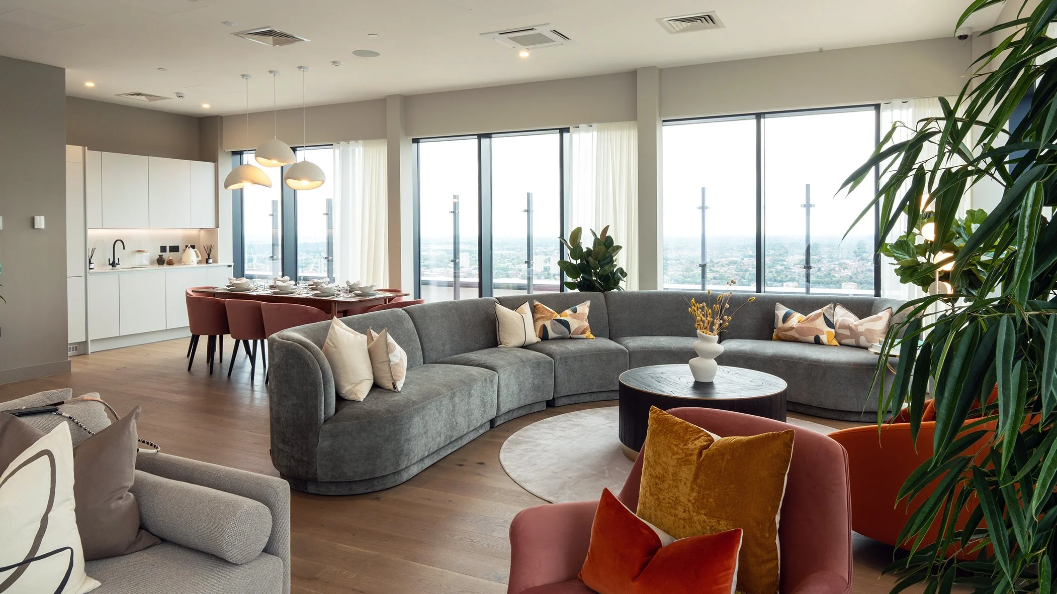 Living room with large windows, gray curved sofa with pillows, round coffee table with a white vase, colorful chairs, a dining table with chairs, white kitchen cabinets, pendant lights, and inside plants.