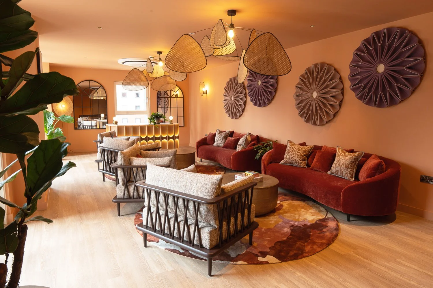 A stylish lounge with two red velvet sofas, beige armchairs, patterned throw pillows, a colorful round area rug, decorative wall art, and a unique light fixture with hanging paper shades. There is a bar counter at the back with a plant and tall mirro